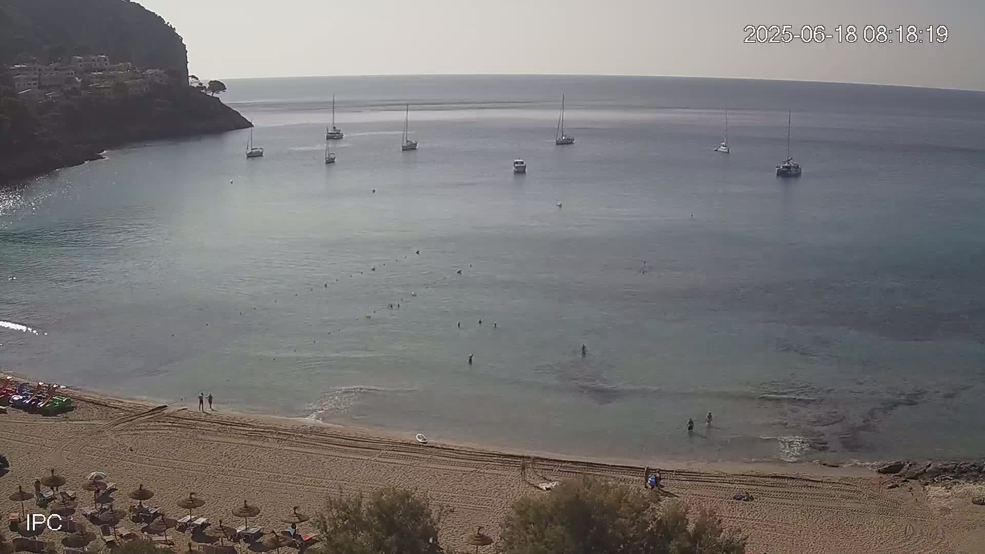 A sandy beach with several people swimming and sunbathing in calm, clear water, with several sailboats anchored in a bay, and a hillside village visible in the background under a hazy sky.