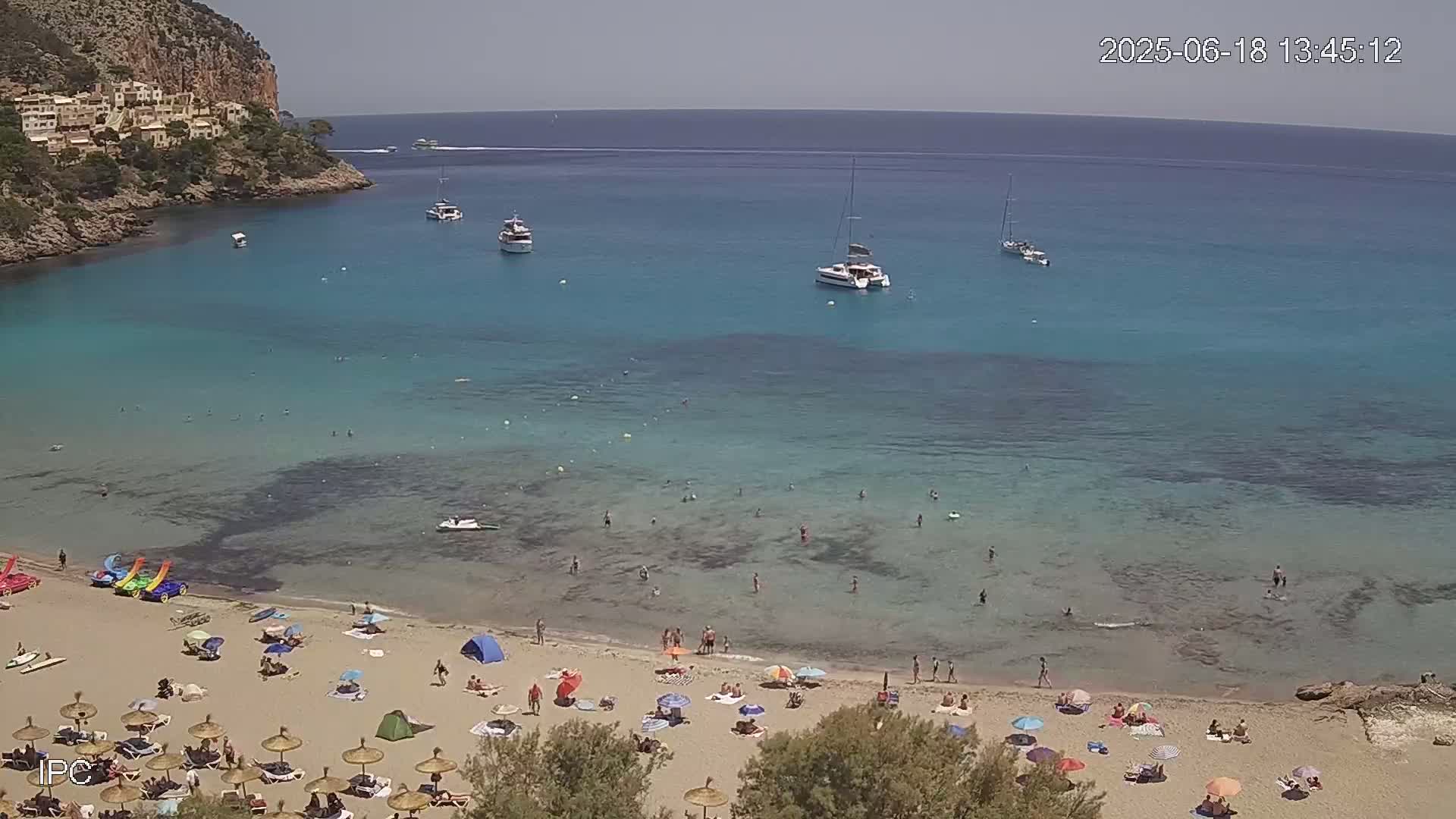 A sunny day at a secluded beach cove shows clear turquoise water with several boats anchored offshore, and people swimming and sunbathing on the sandy shore.