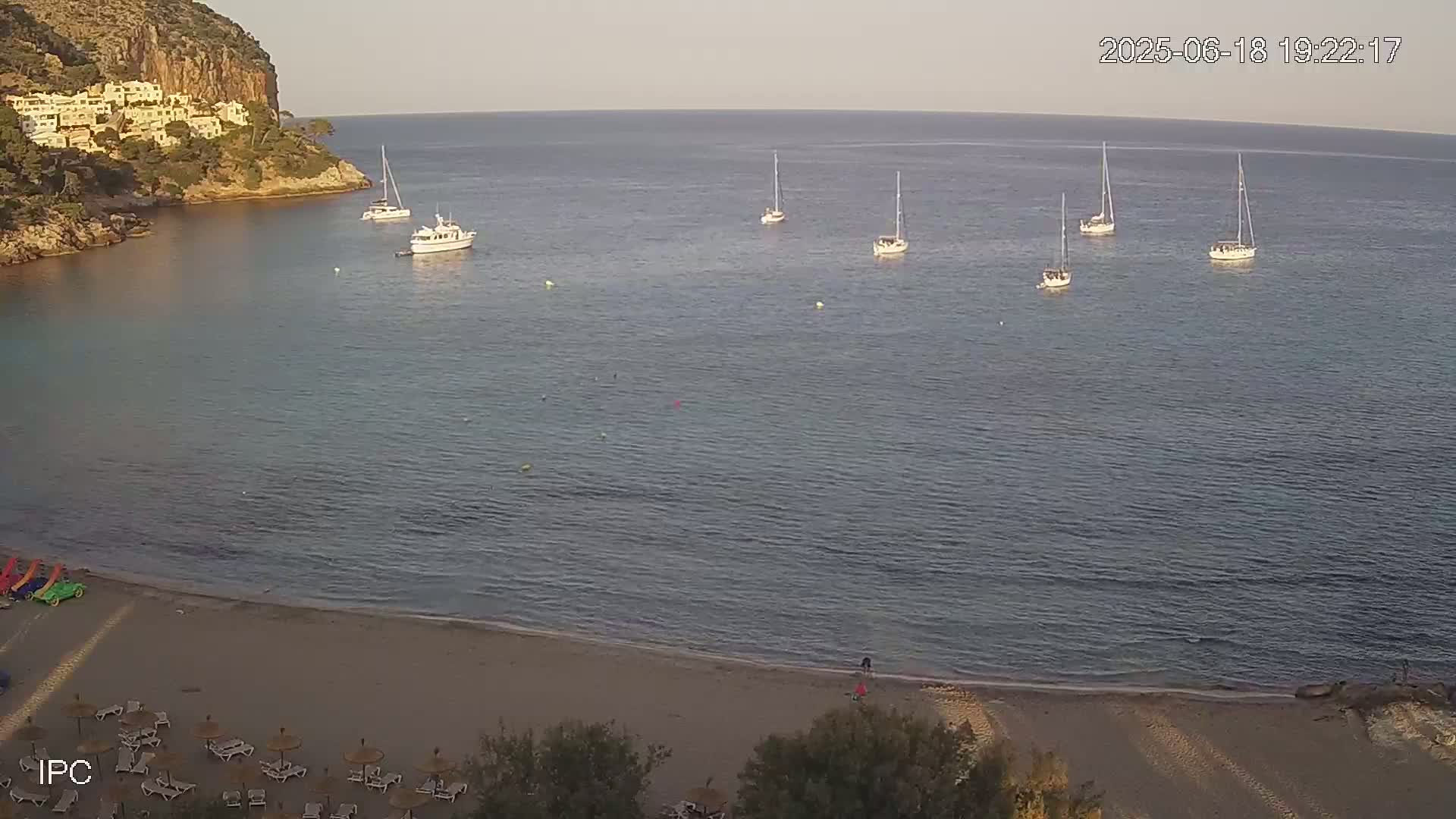 A sandy beach with lounge chairs and umbrellas sits below a calm bay where several sailboats are anchored near a rocky cliff with buildings on top.