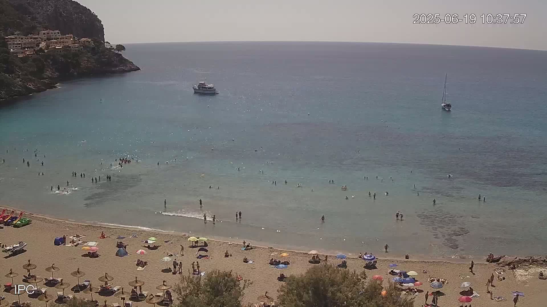 A sandy beach with numerous people and umbrellas is nestled in a cove with clear, shallow turquoise water, a boat, and a sailboat visible in the distance on a sunny day.
