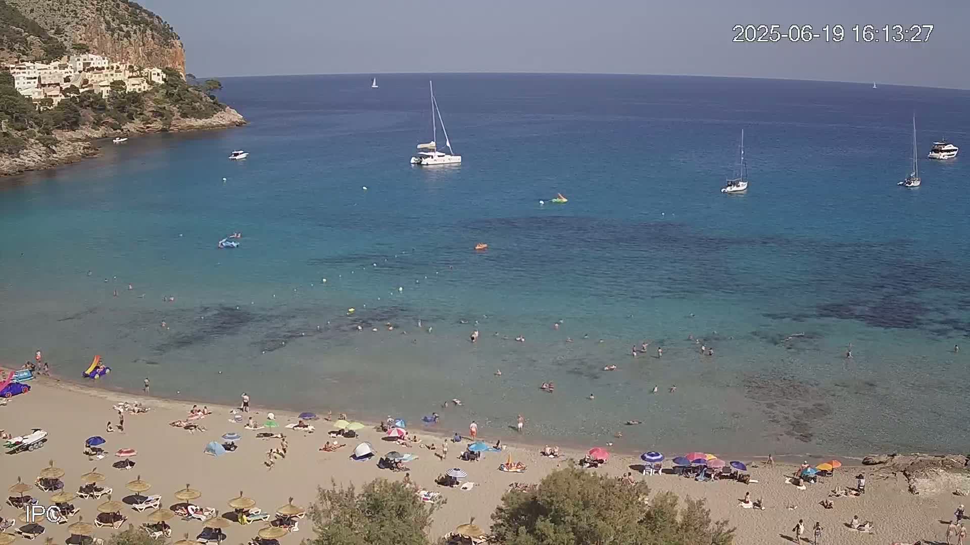 A sandy beach with many people and colorful umbrellas is nestled in a cove with clear turquoise water, several sailboats, and a rocky cliff in the background on a sunny day.