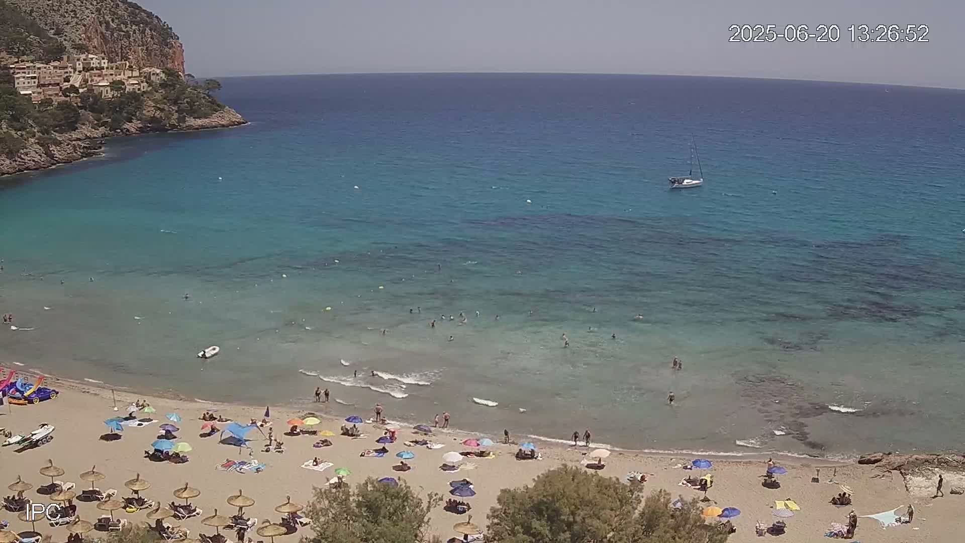 A sandy beach with colorful umbrellas and people swimming in clear, turquoise water, backed by a rocky cliff and a sailboat on a sunny day.