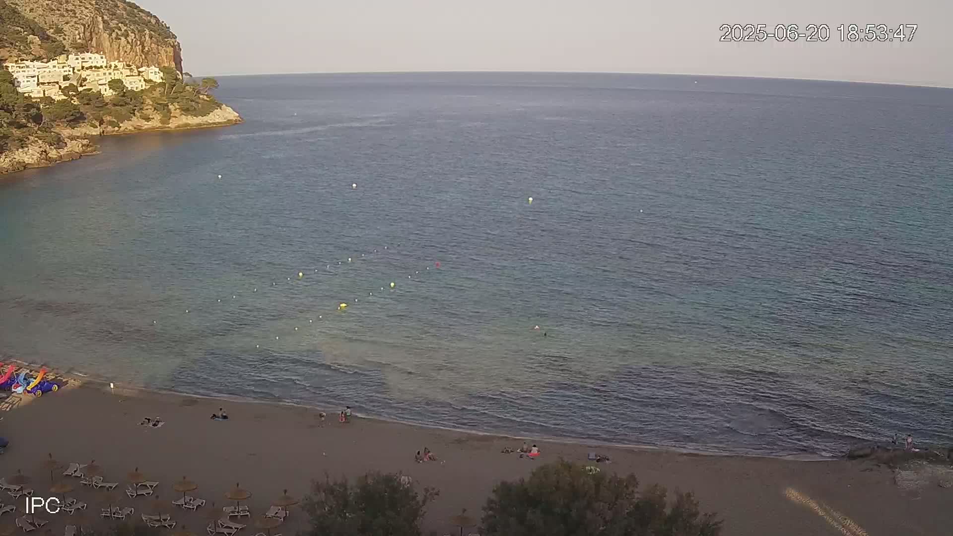 A sandy beach with a few people on it and several lounge chairs under umbrellas is next to a calm, clear ocean, with a rocky cliff and buildings visible in the background under a mostly clear sky.