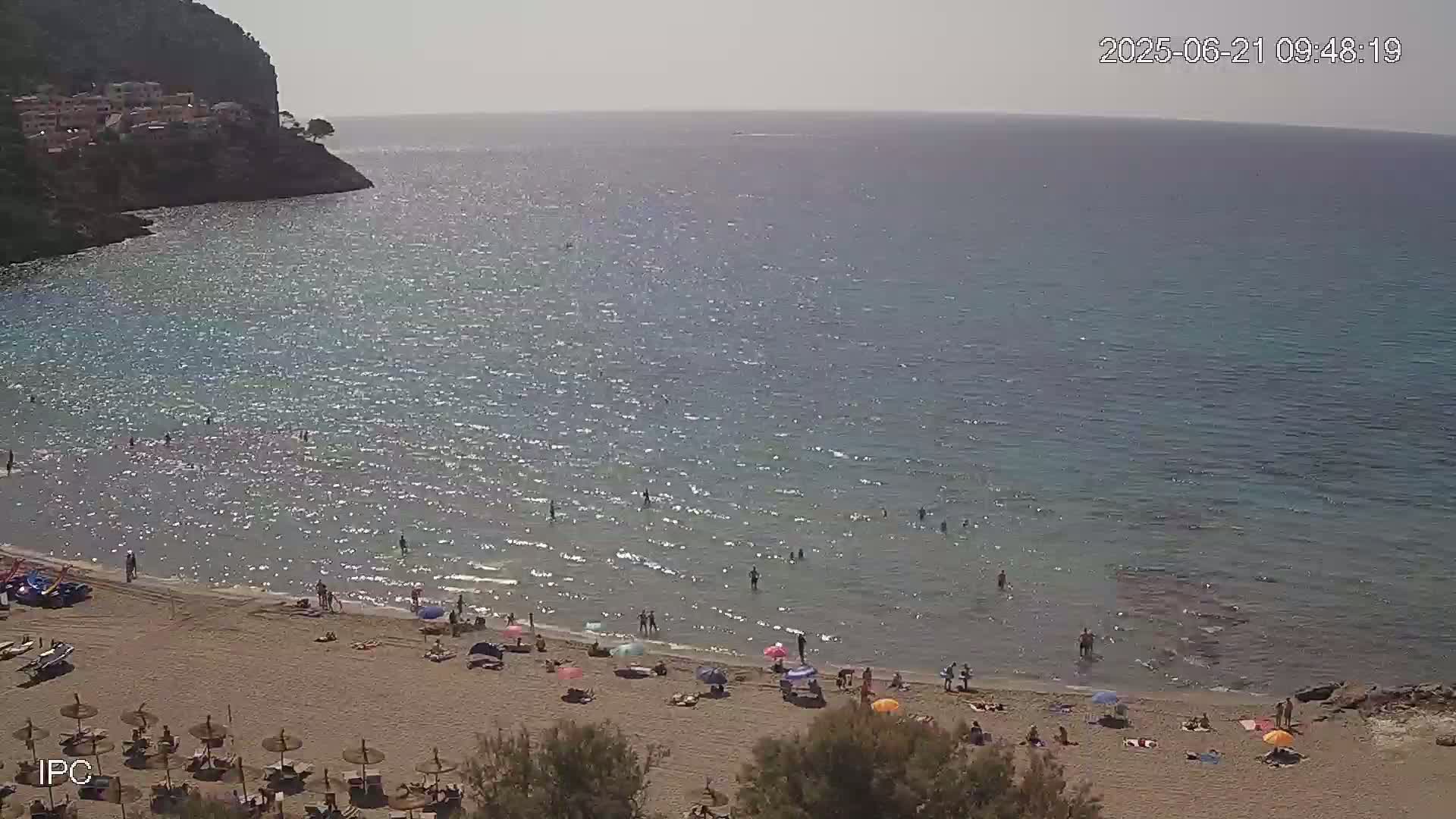 A sandy beach with people swimming and sunbathing under umbrellas on a sunny day, next to a calm, sparkling sea and a rocky cliff.