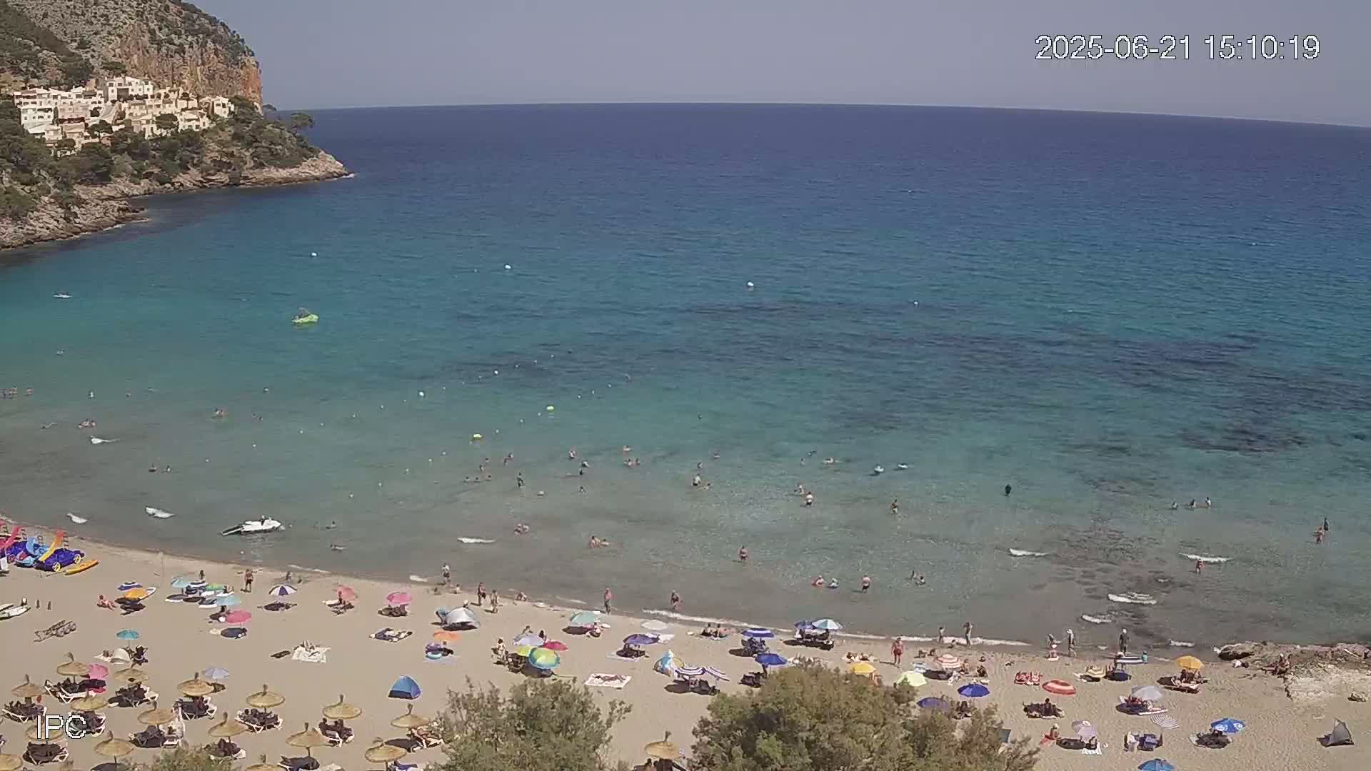 A sandy beach with numerous umbrellas and people swimming in clear, shallow turquoise water, backed by a rocky cliff on a sunny day.