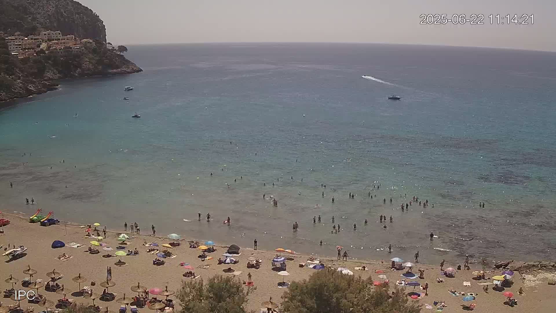 A sandy beach with numerous people swimming and sunbathing under umbrellas, situated below a rocky cliff face on a sunny day, overlooks a calm, clear ocean with several small boats.