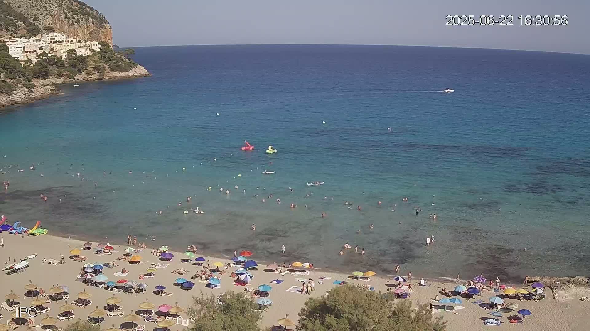 A sandy beach with numerous colorful umbrellas and sunbathers is situated on a clear, turquoise bay next to a rocky cliff on a sunny day.