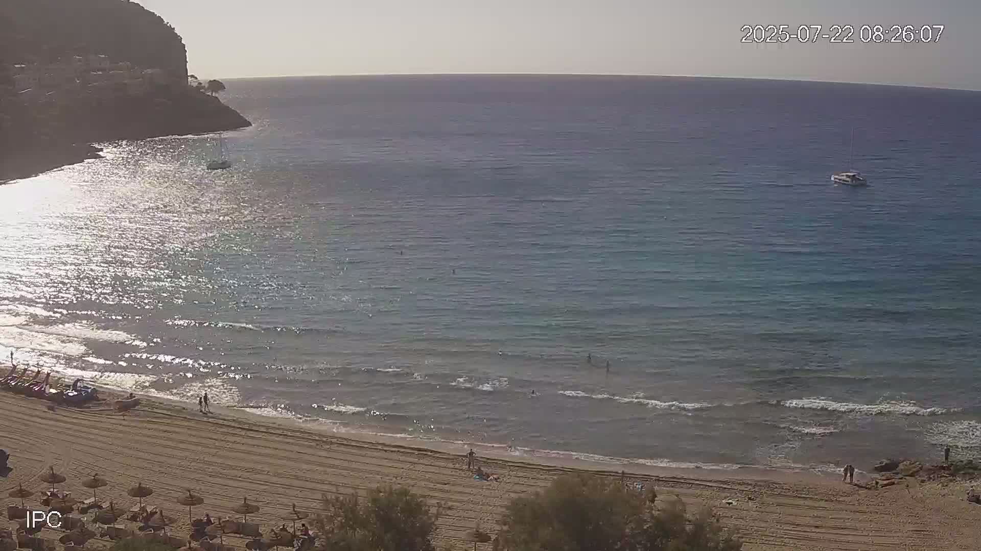 A sandy beach with calm, clear water laps the shore under a bright, sunny sky, with a few people swimming and sunbathing, and a sailboat visible in the distance.
