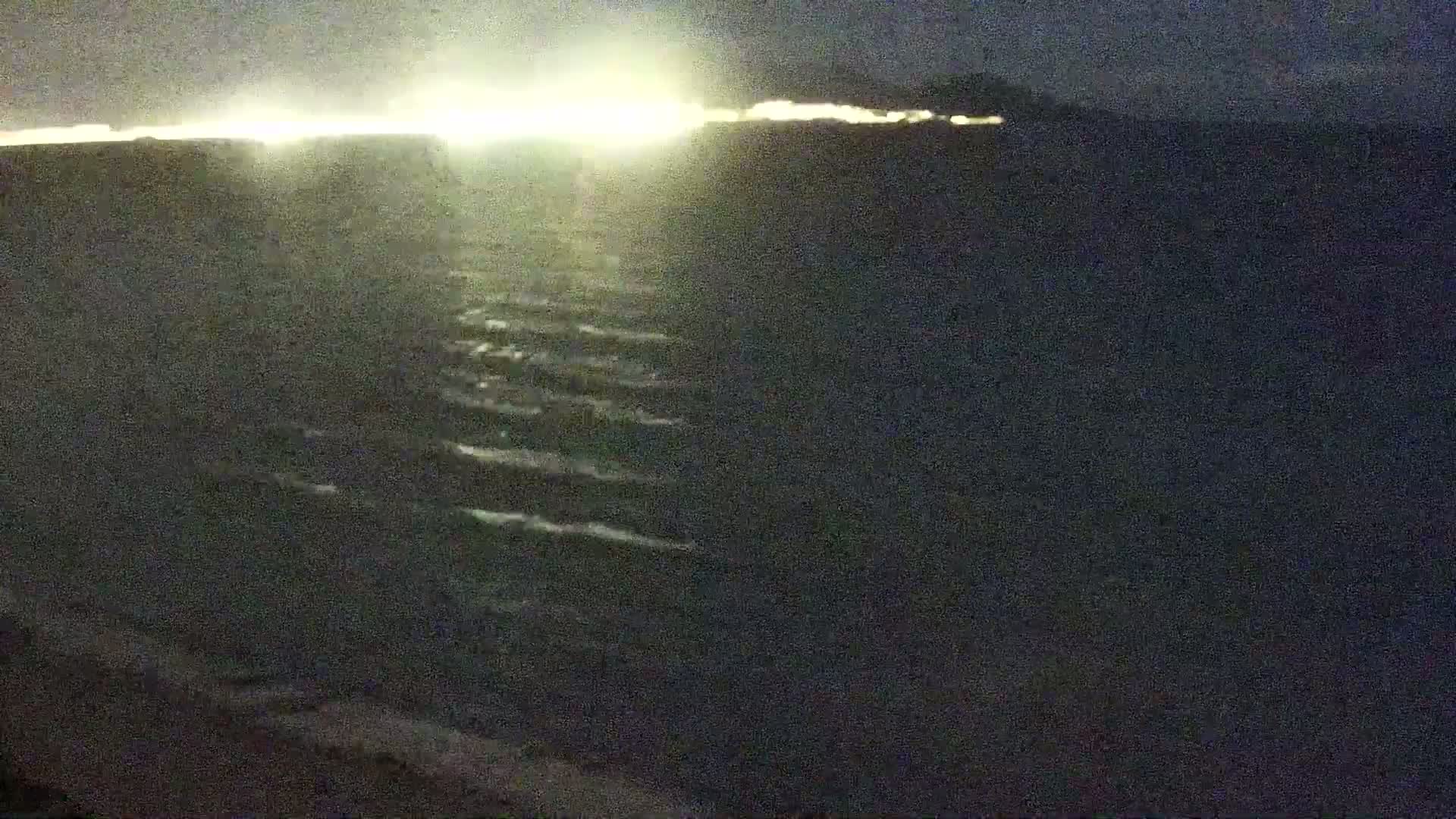 A dark body of water with small waves is visible under a nighttime sky illuminated by a bright light source on the horizon.