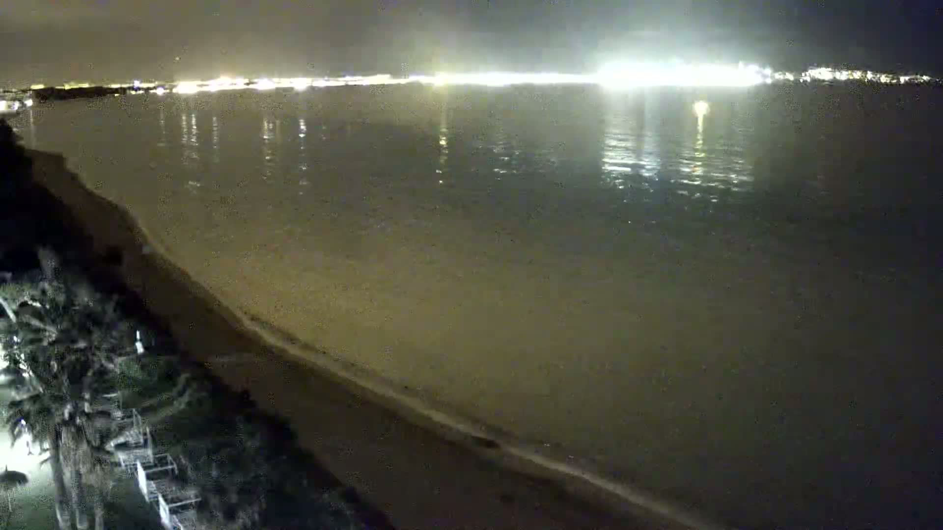 A nighttime view shows a calm body of water reflecting city lights across its surface, with a dark sandy beach and vegetation visible in the foreground.