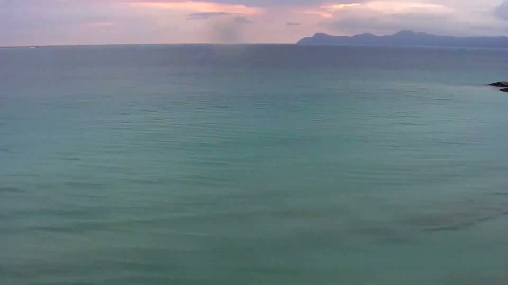 A calm, teal ocean stretches to a distant, mountainous shoreline under a cloudy, pinkish sky.