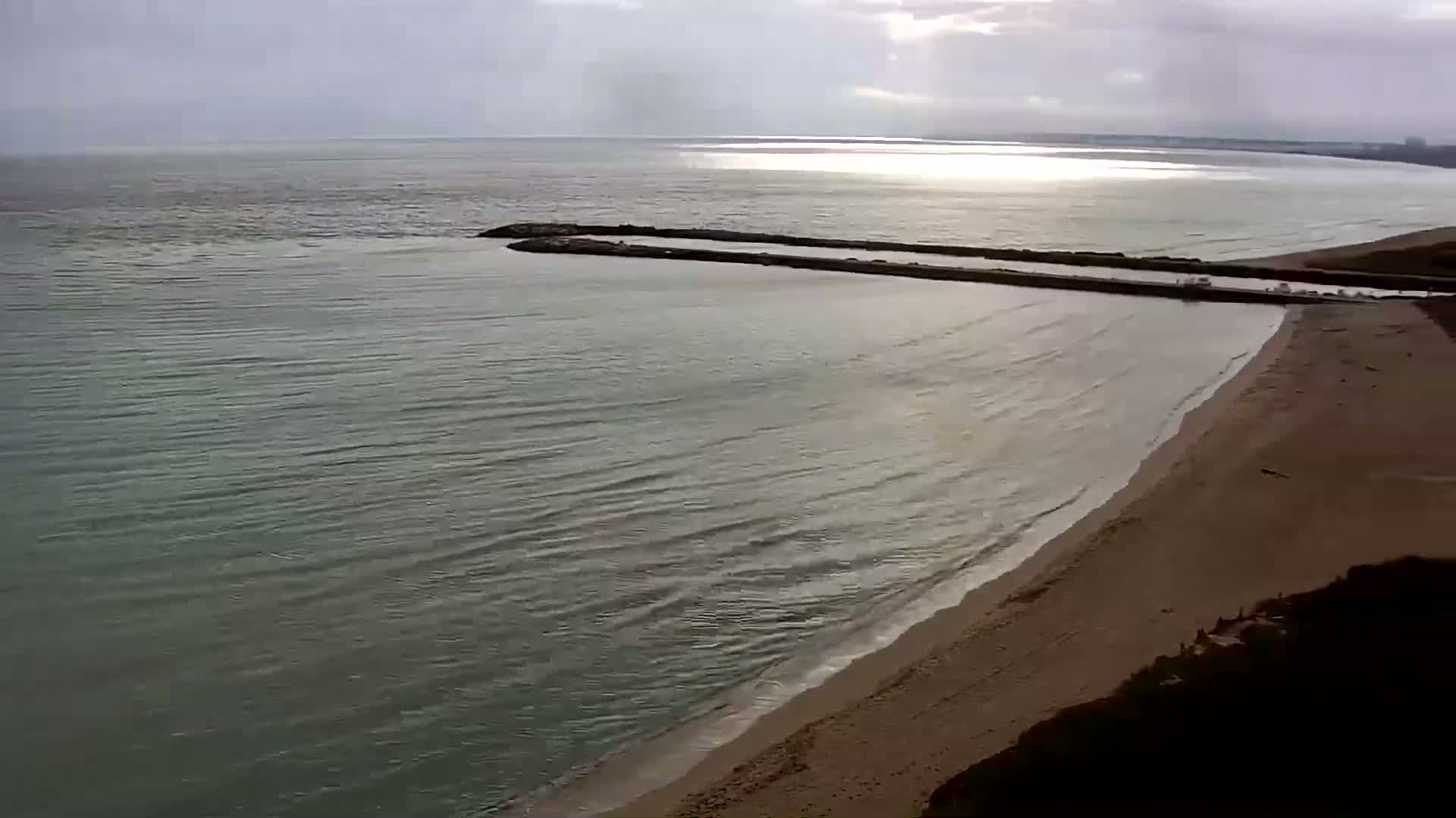 A sandy beach meets calm, green-gray water under a cloudy sky, with two long jetties extending into the sea.