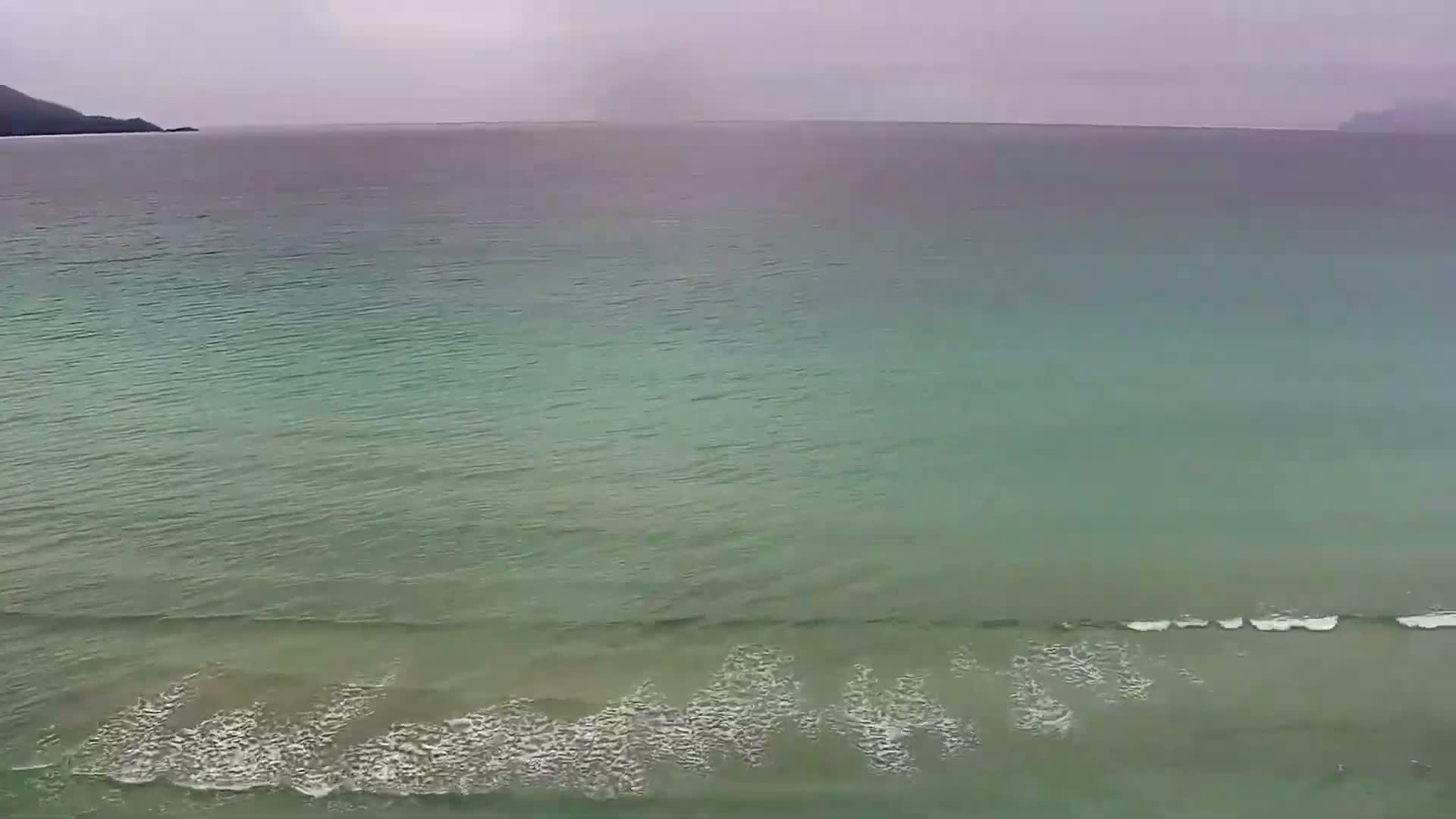 A tranquil, teal-colored ocean with small waves laps a shore under an overcast sky.