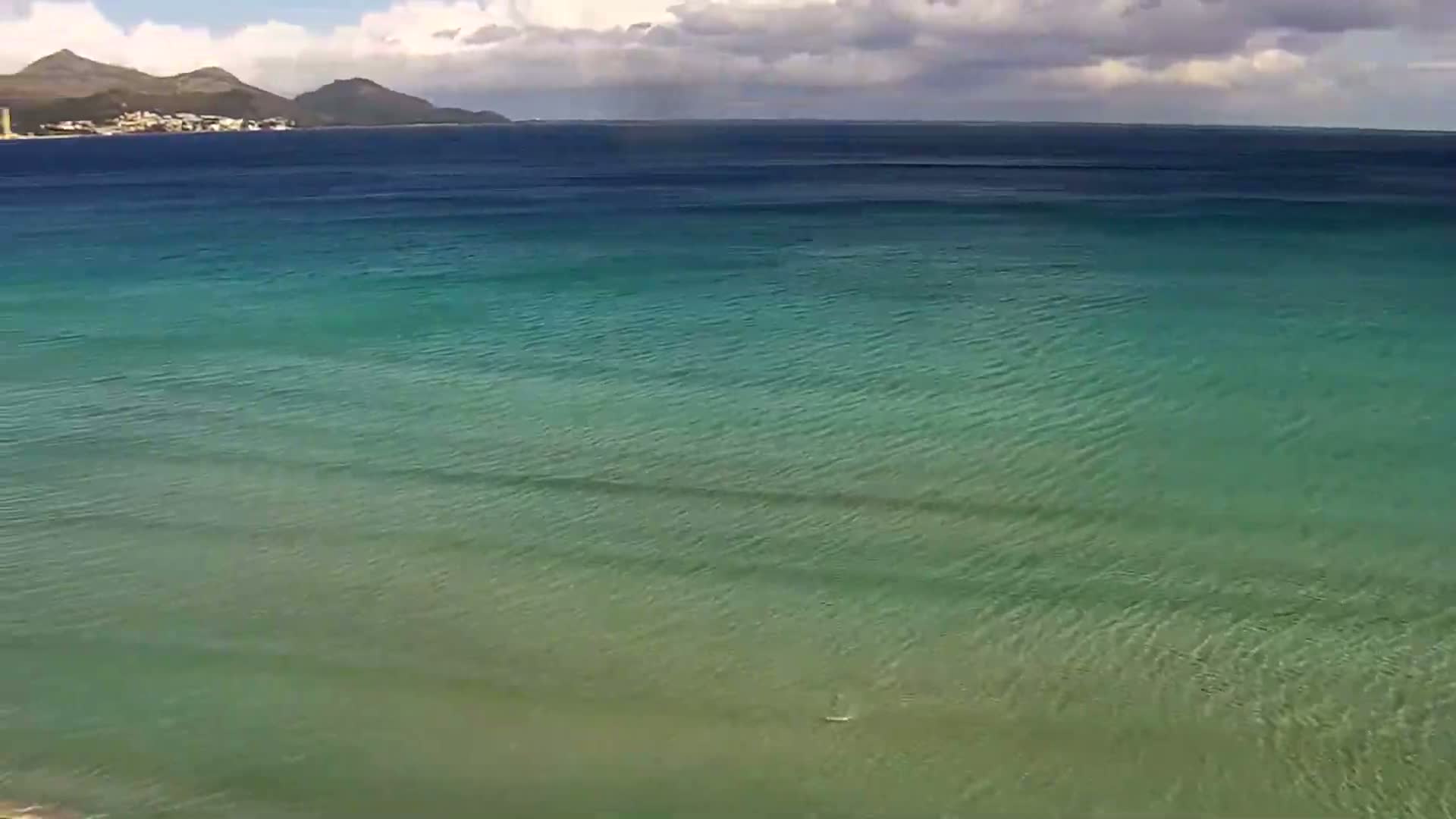 A calm, teal ocean stretches to a distant shoreline under a partly cloudy sky.