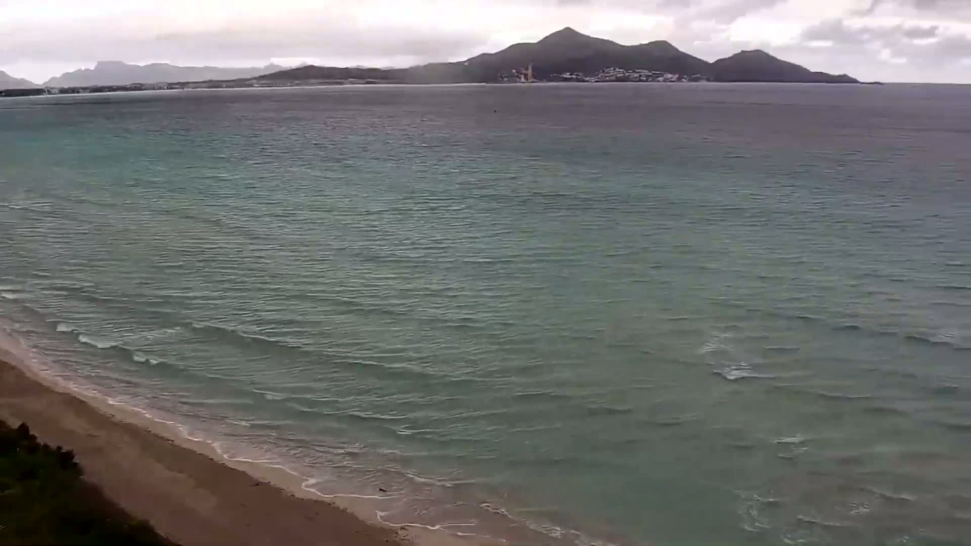 A sandy beach meets a calm, greenish-grey sea under an overcast sky, with a distant mountain range visible on the horizon.