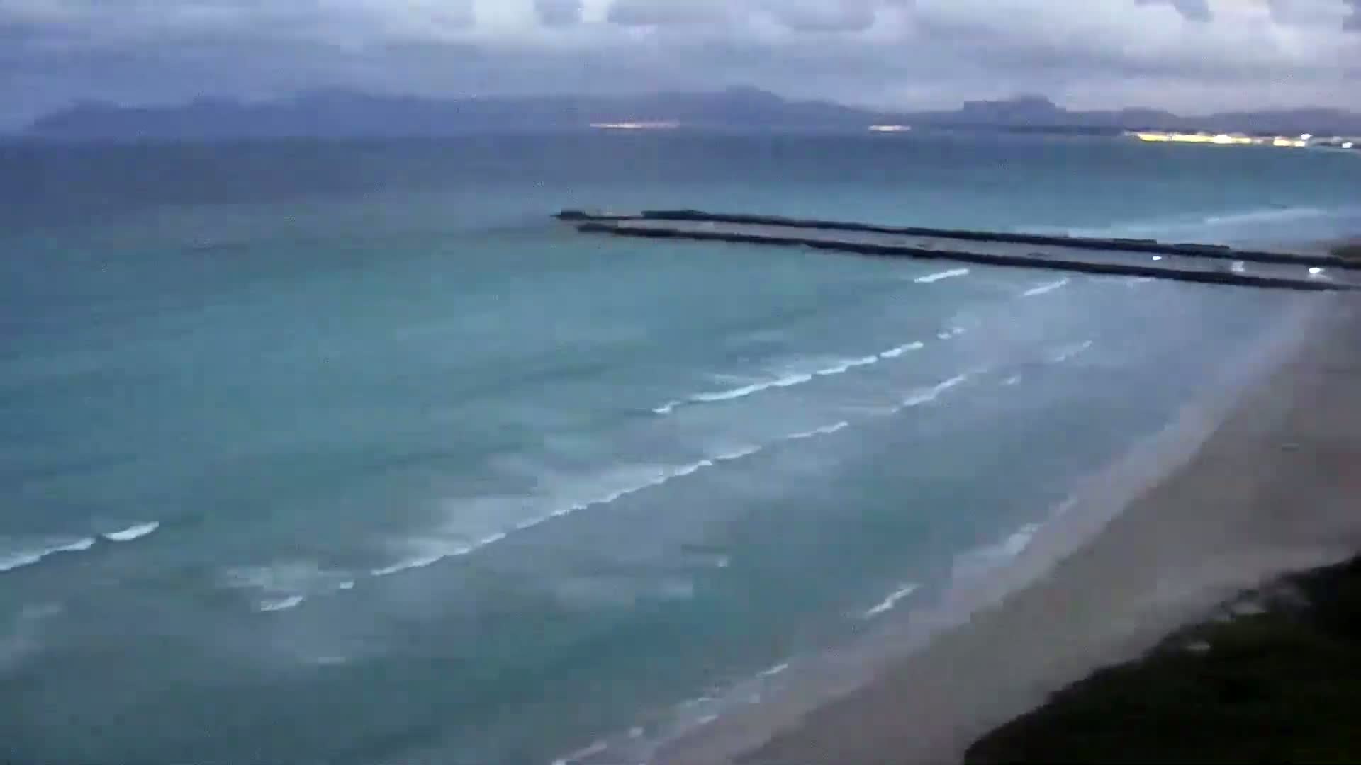 A long, dark jetty extends into a teal ocean with small waves lapping a sandy beach under an overcast sky.