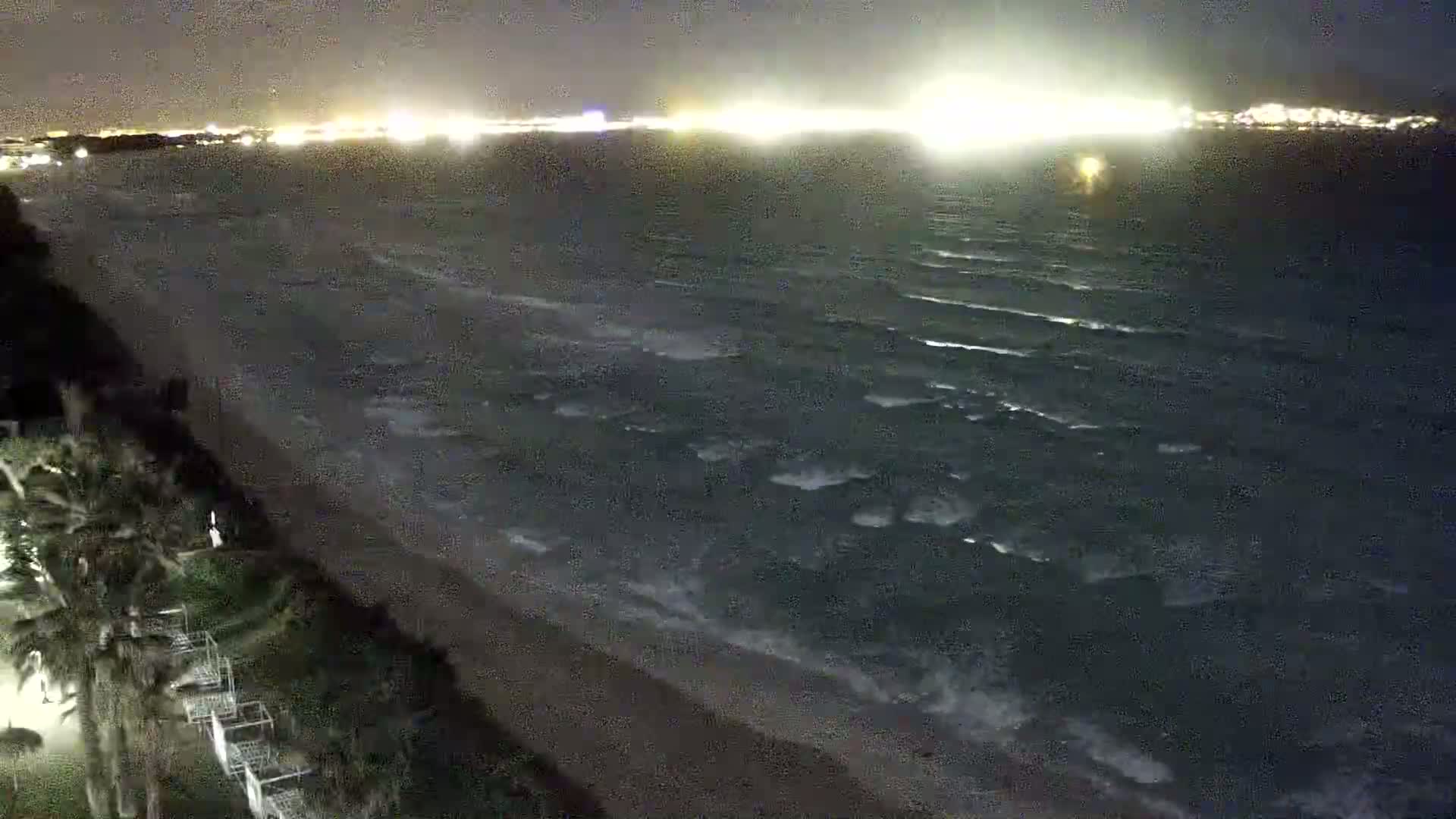 A night-time view of a wavy ocean meeting a sandy shore lined with vegetation under a dark sky illuminated by distant city lights.