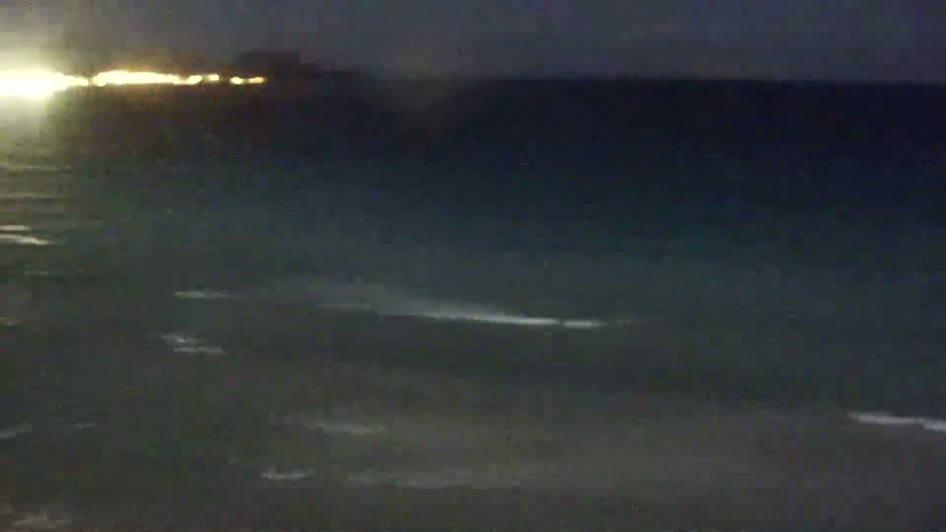A dark, nighttime view of a calm body of water with a distant line of lights visible on the horizon.