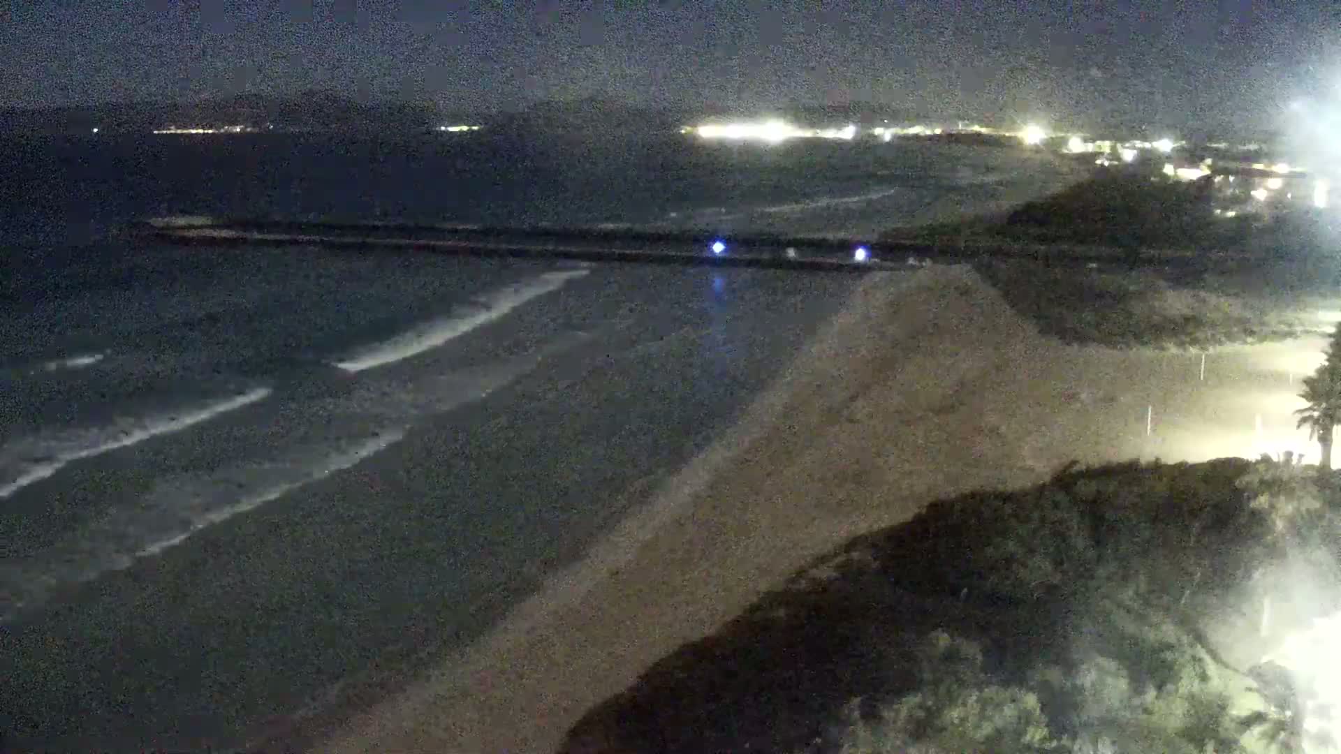 At night, gentle waves lap a sandy beach, with a long jetty extending into the dark ocean under a clear sky, and distant lights illuminating the coastline.