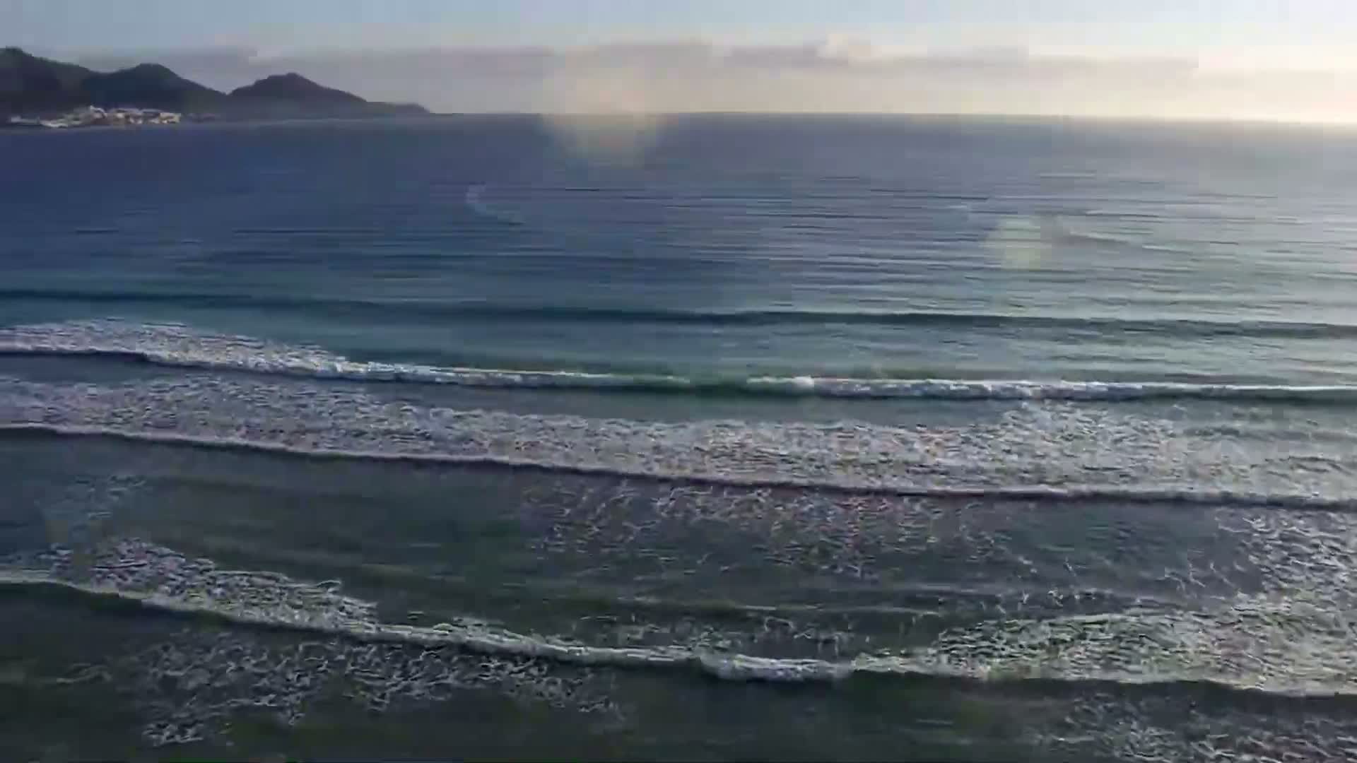 Gentle waves roll onto a calm ocean shore under a mostly clear sky, with distant land visible on the horizon.
