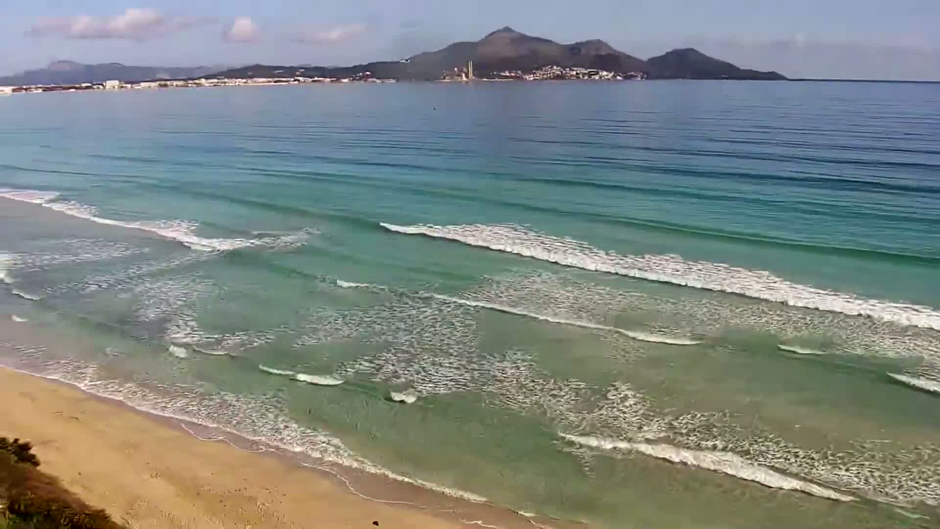 Gentle waves roll onto a sandy beach under a partly cloudy sky, with a distant mountain visible across the calm water.