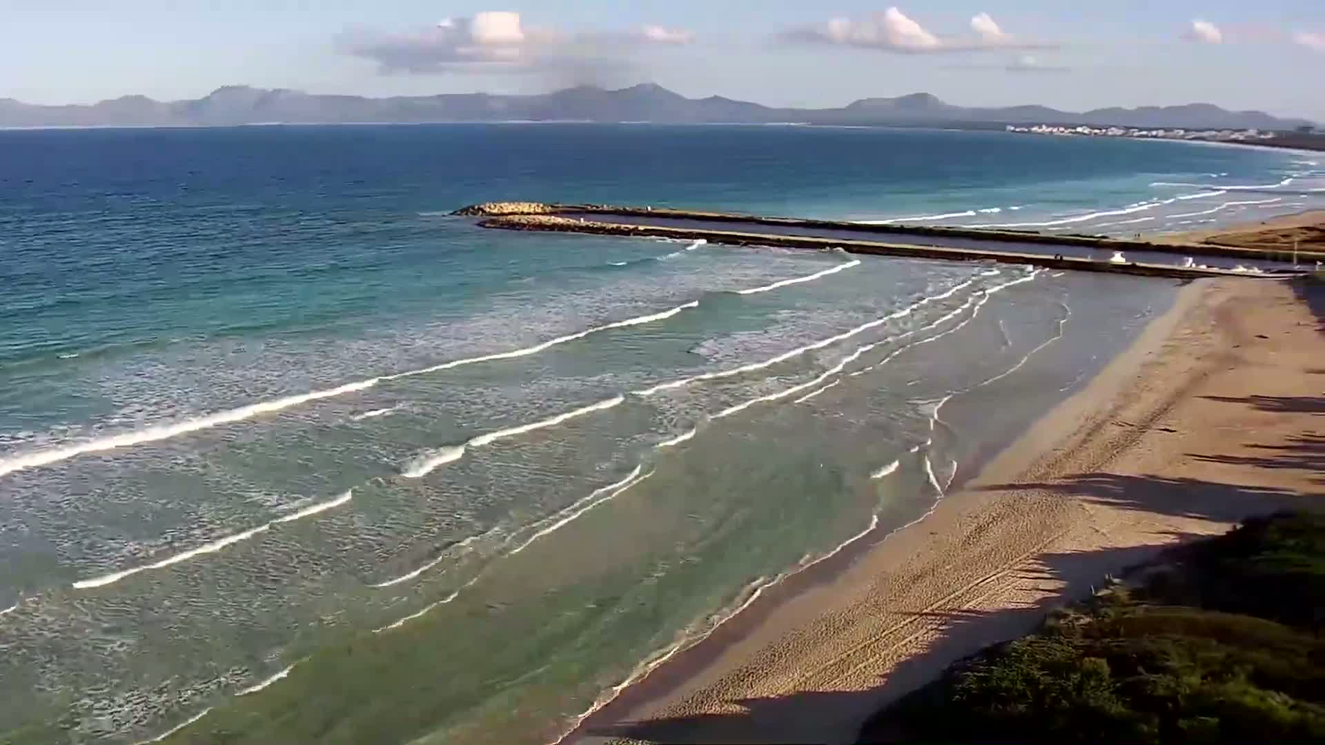A sandy beach with gentle waves rolling onto the shore under a partly cloudy sky, a breakwater extending into the ocean, and mountains visible in the distance.