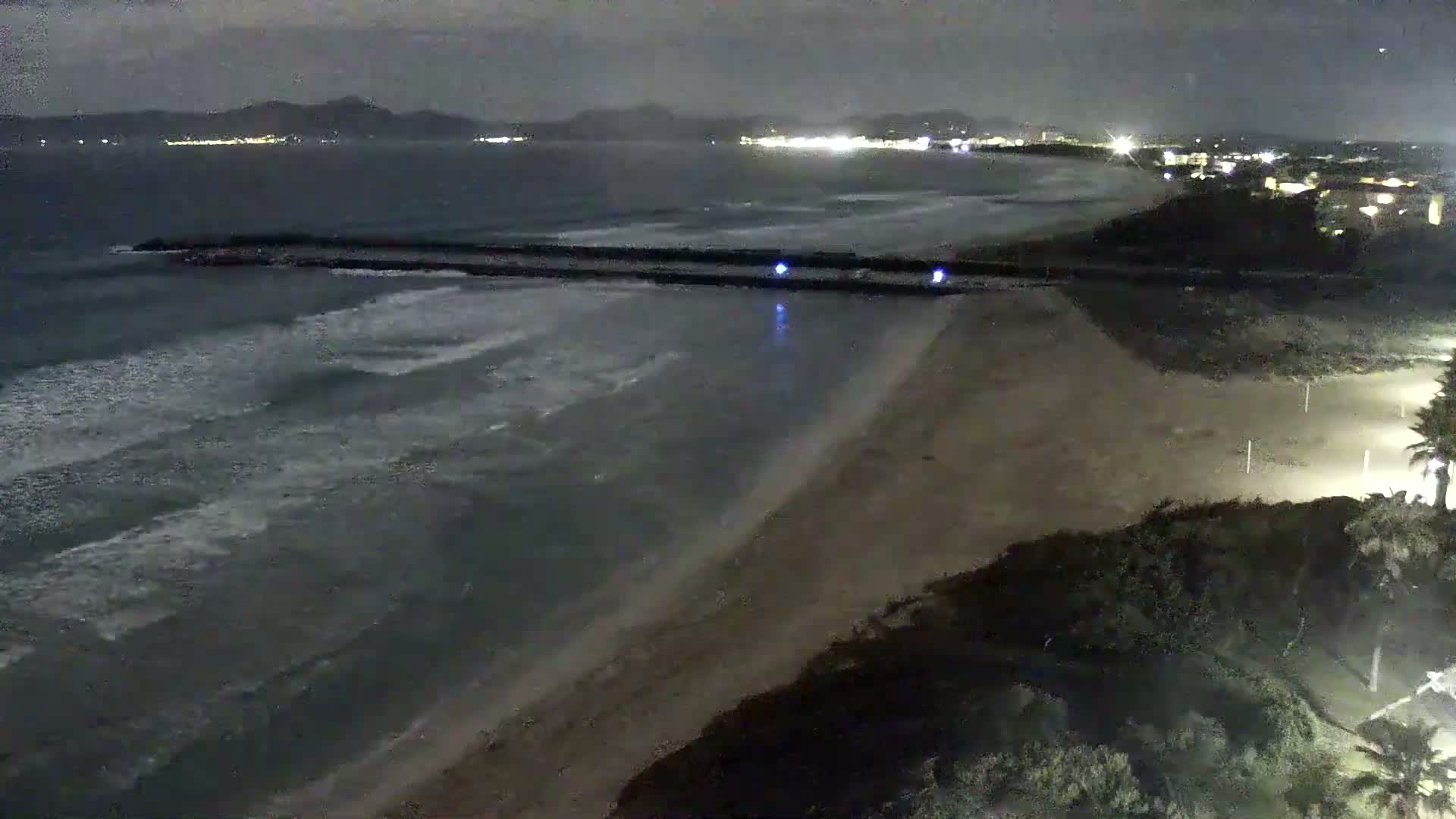 A dark, nighttime view of a beach with gentle waves lapping the shore, under a cloudy sky, with lights visible in the distance on land.