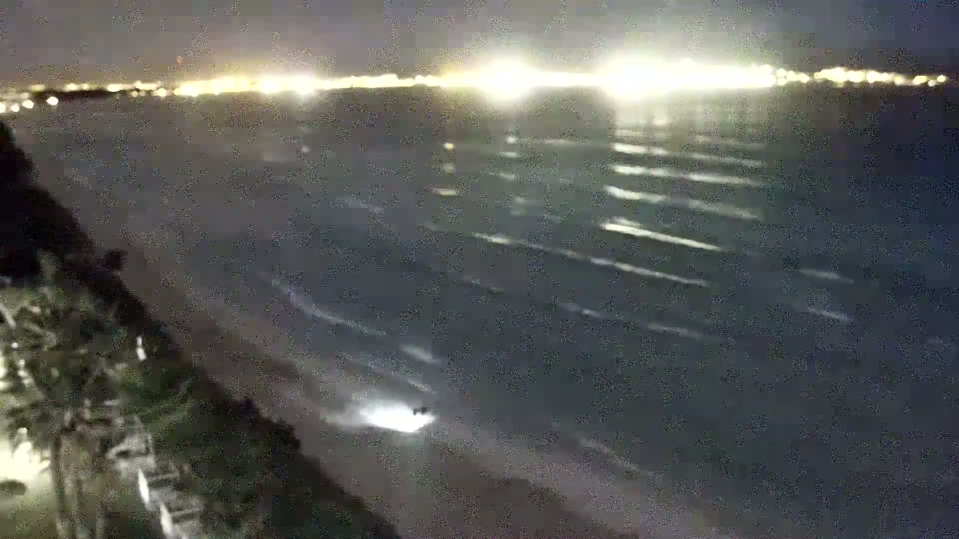 A nighttime view shows a dark beach with gentle waves lapping the shore under a clear sky, with city lights visible in the distance.