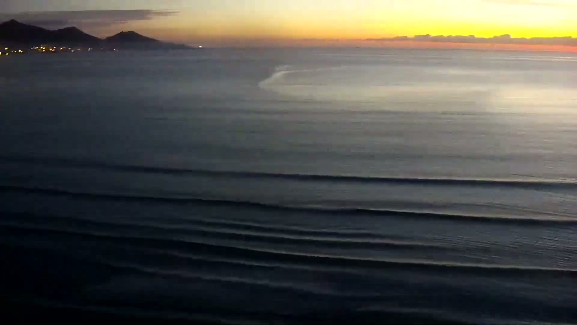 A hazy sunrise illuminates a calm ocean with small waves rolling toward a distant, dimly lit shoreline beneath a mostly clear sky.