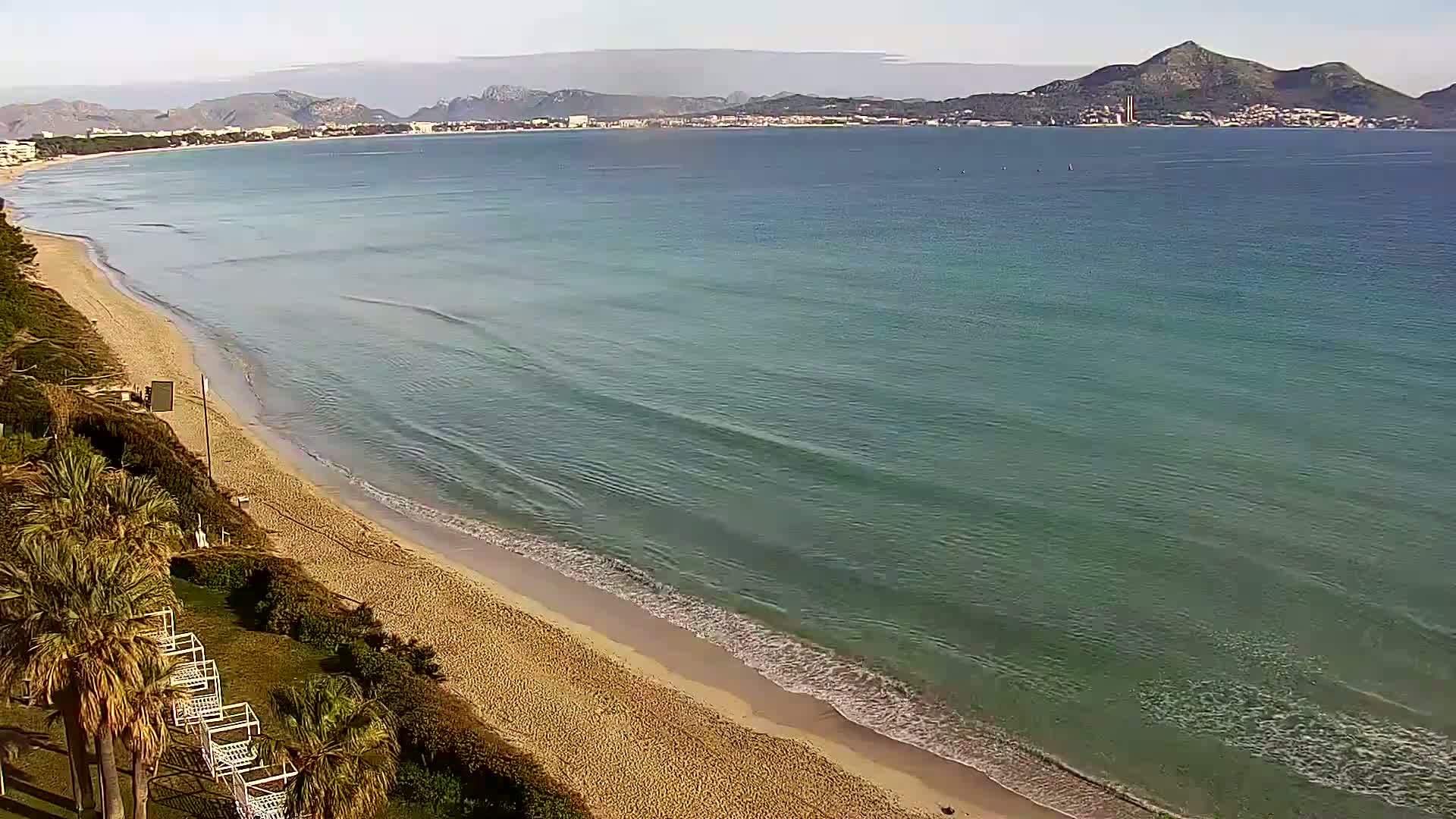 A sandy beach with palm trees and gentle waves laps the shore under a clear sky, with mountains visible in the distance.