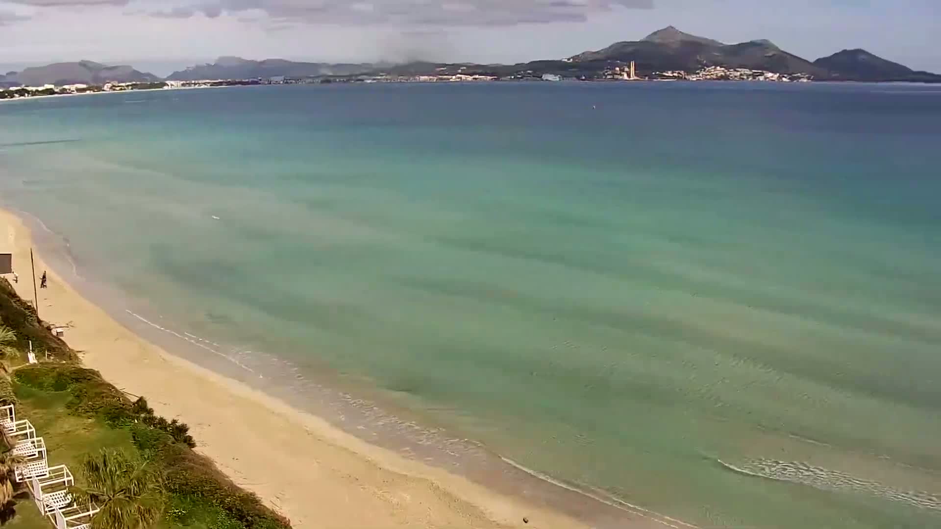 A sandy beach slopes gently into a calm, teal-colored sea, with a distant mountain range visible under a partly cloudy sky.