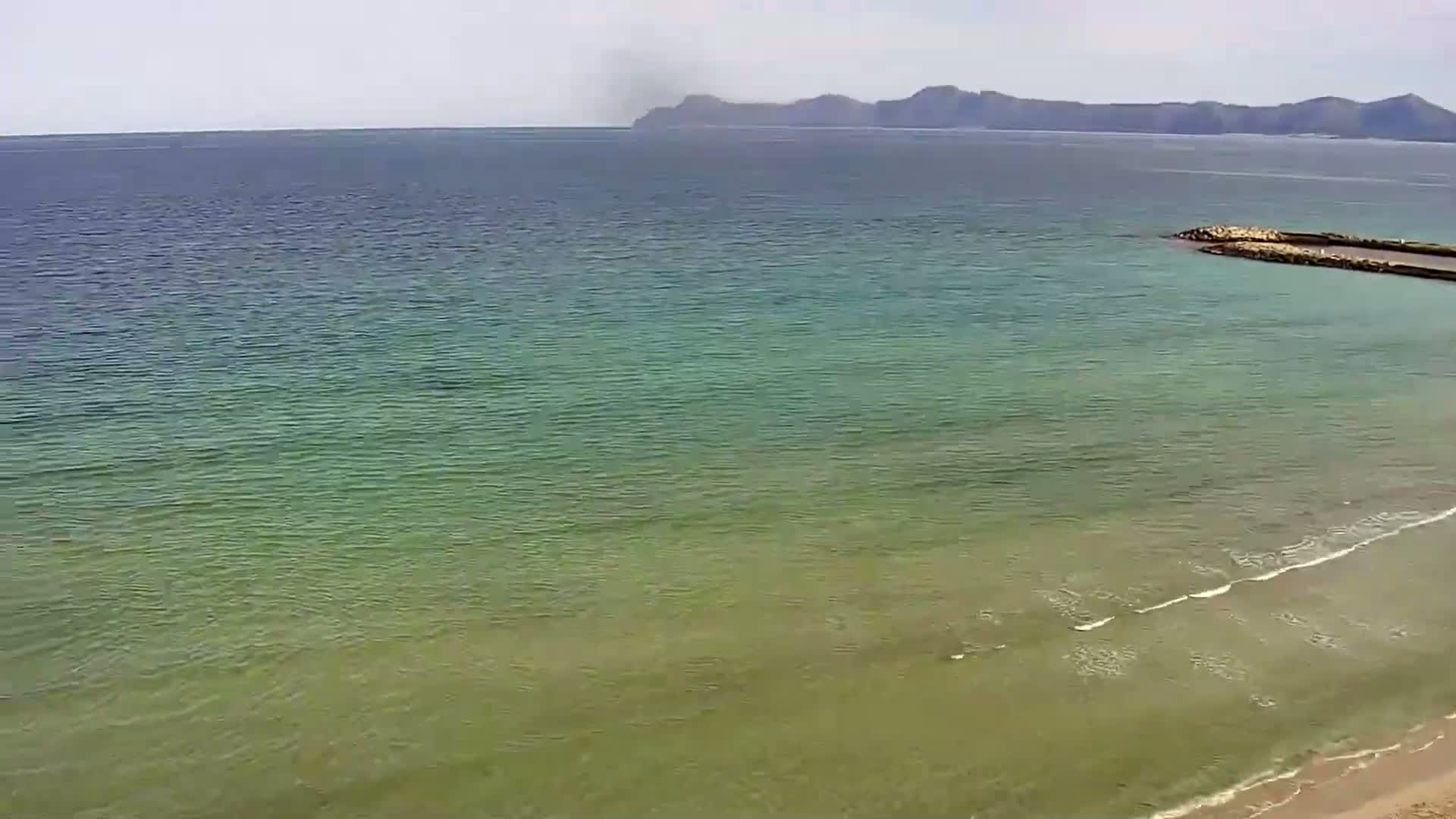 A calm, green-tinged sea stretches to a distant mountain range under a hazy sky, with a small breakwater visible near the shore.