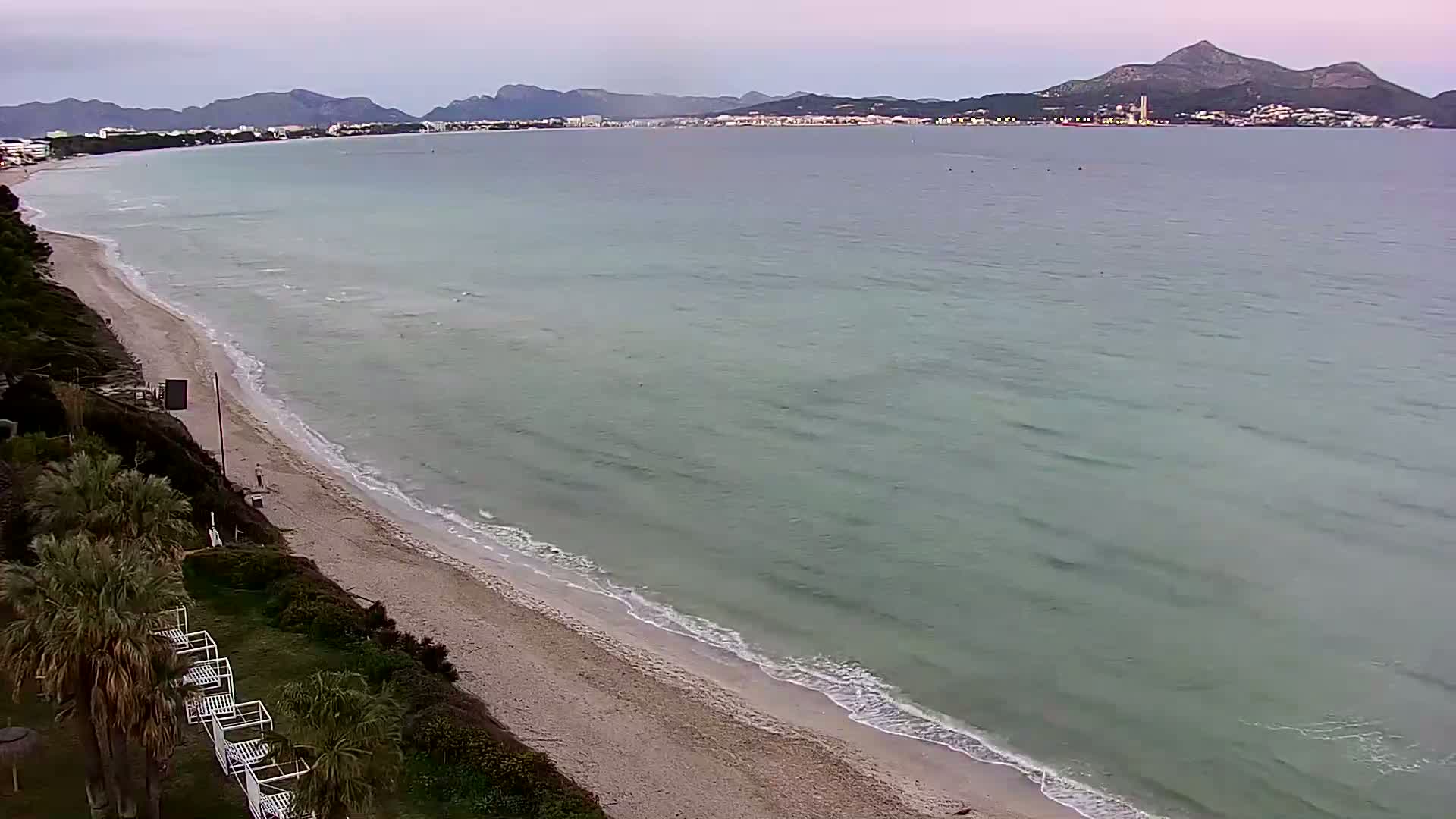 An aerial view shows a sandy beach with gentle waves lapping the shore under an overcast sky, with mountains visible in the distance.