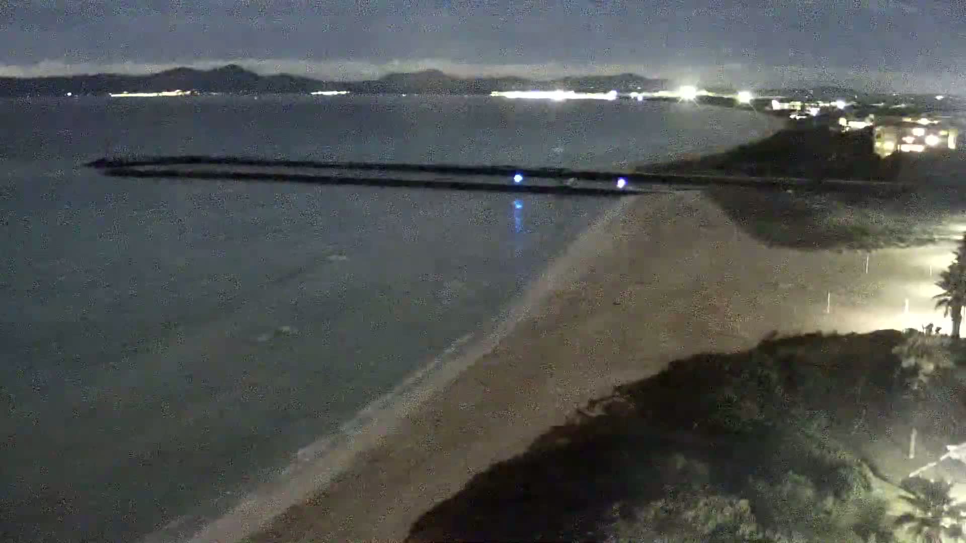 A nighttime view of a sandy beach and calm ocean with a long, dark breakwater extending into the water, under a dark sky.