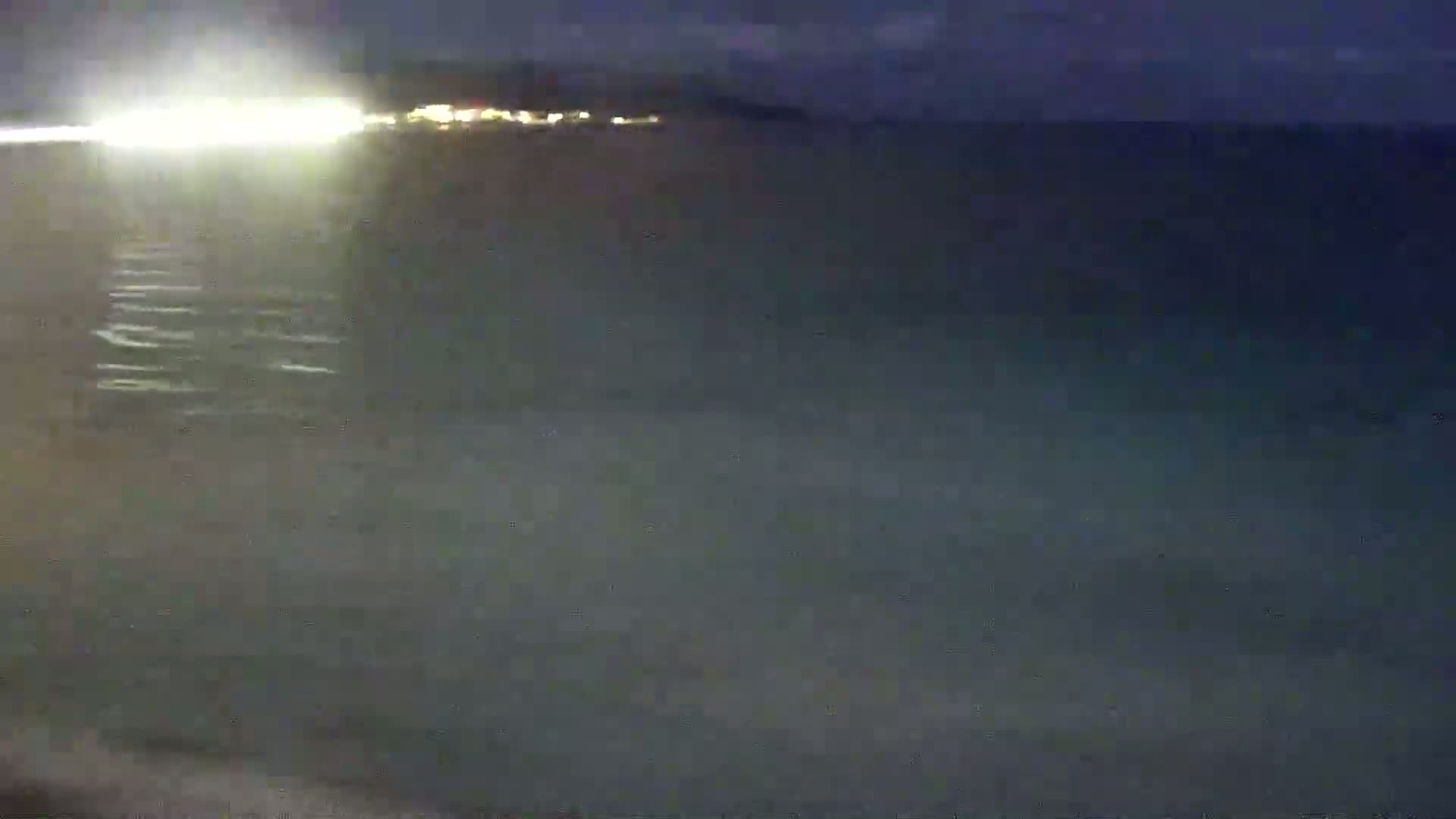 A dark, calm body of water reflects a distant, brightly lit shoreline at night.