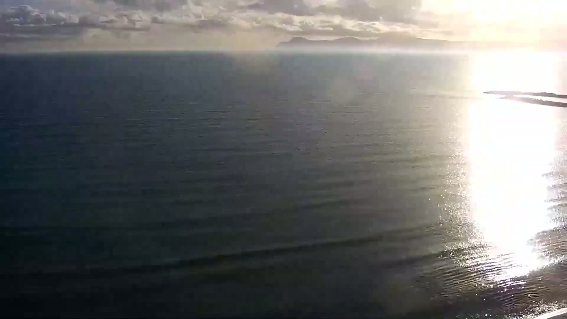 A calm sea reflects sunlight under a partly cloudy sky, with a distant landmass visible on the horizon.
