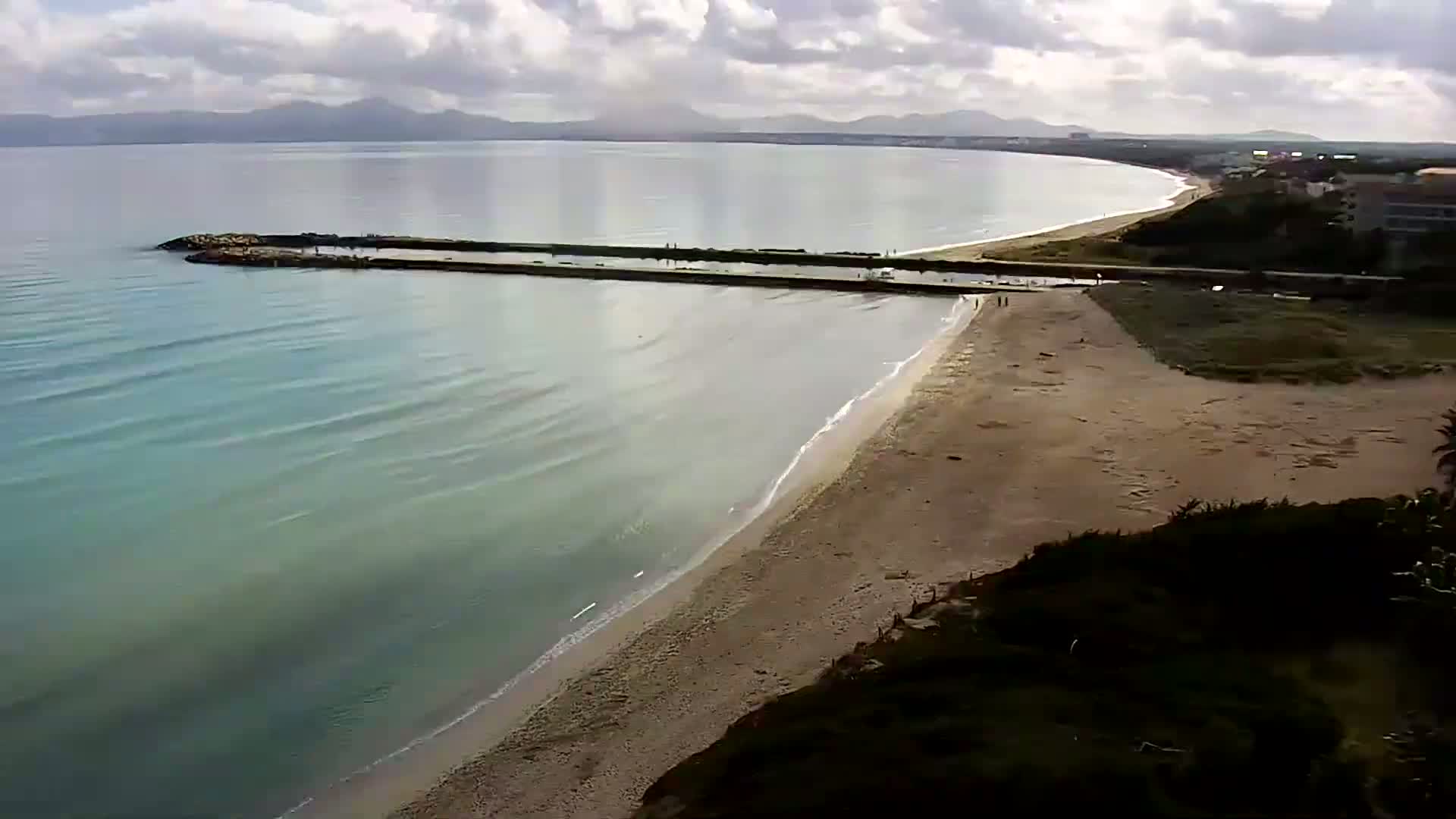 A sandy beach with calm, light-blue water meets a long, low-lying breakwater, with mountains visible in the distance under a partly cloudy sky.