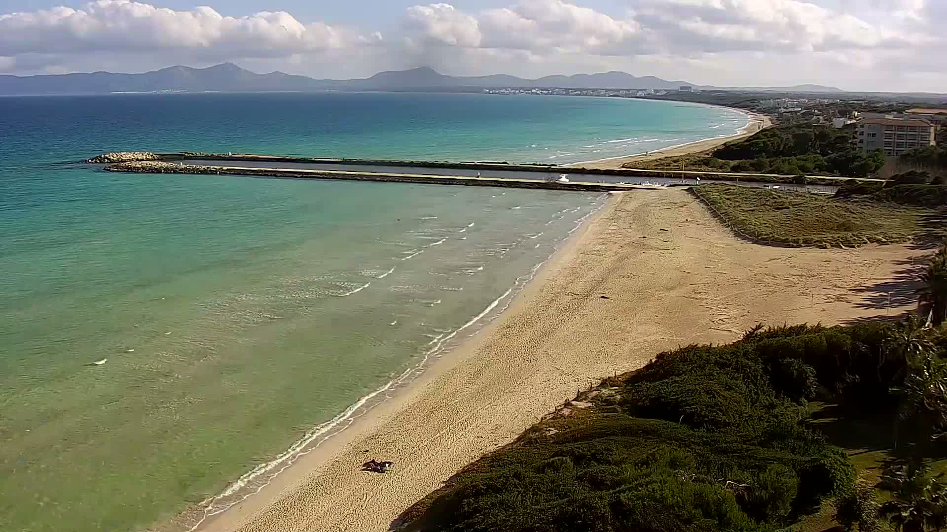 A sandy beach with calm, clear turquoise water stretches along a coastline under a partly cloudy sky, with mountains visible in the distance.