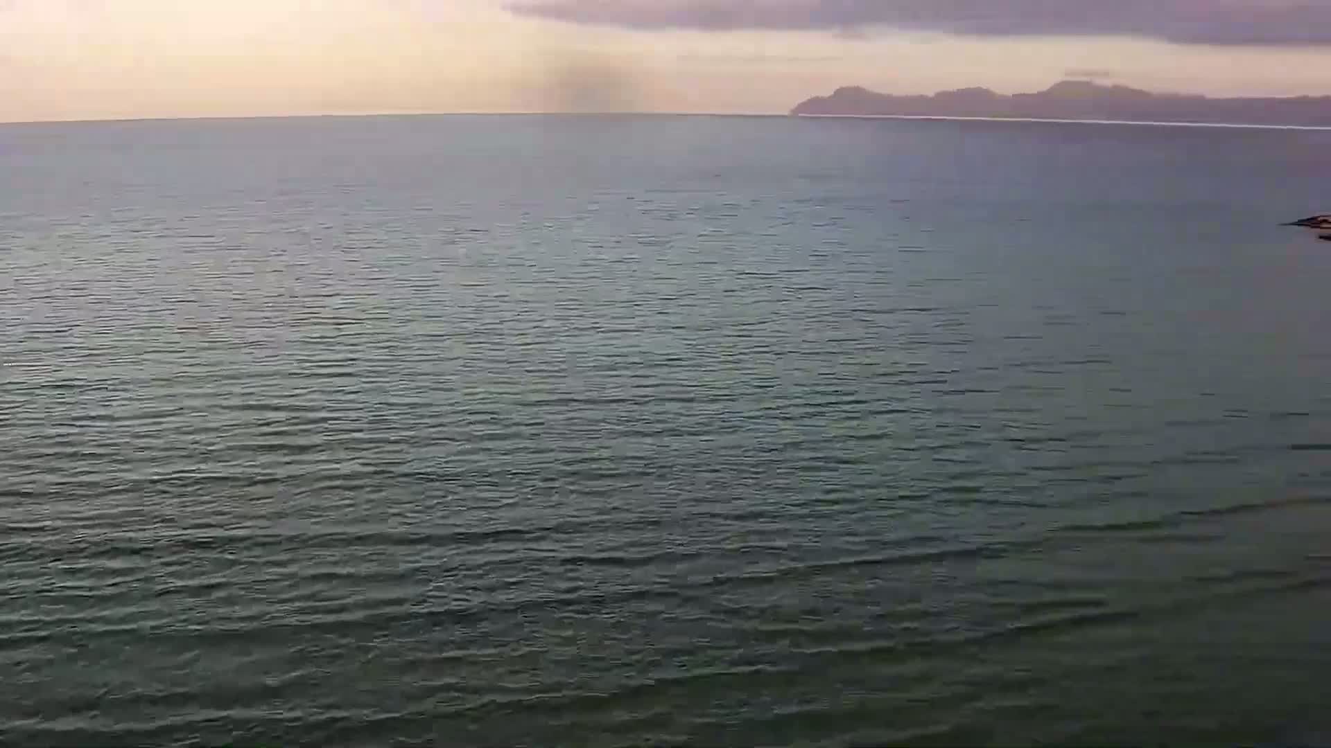 A calm, dark-grey sea stretches to a distant, hazy mountain range under a mostly cloudy sky.