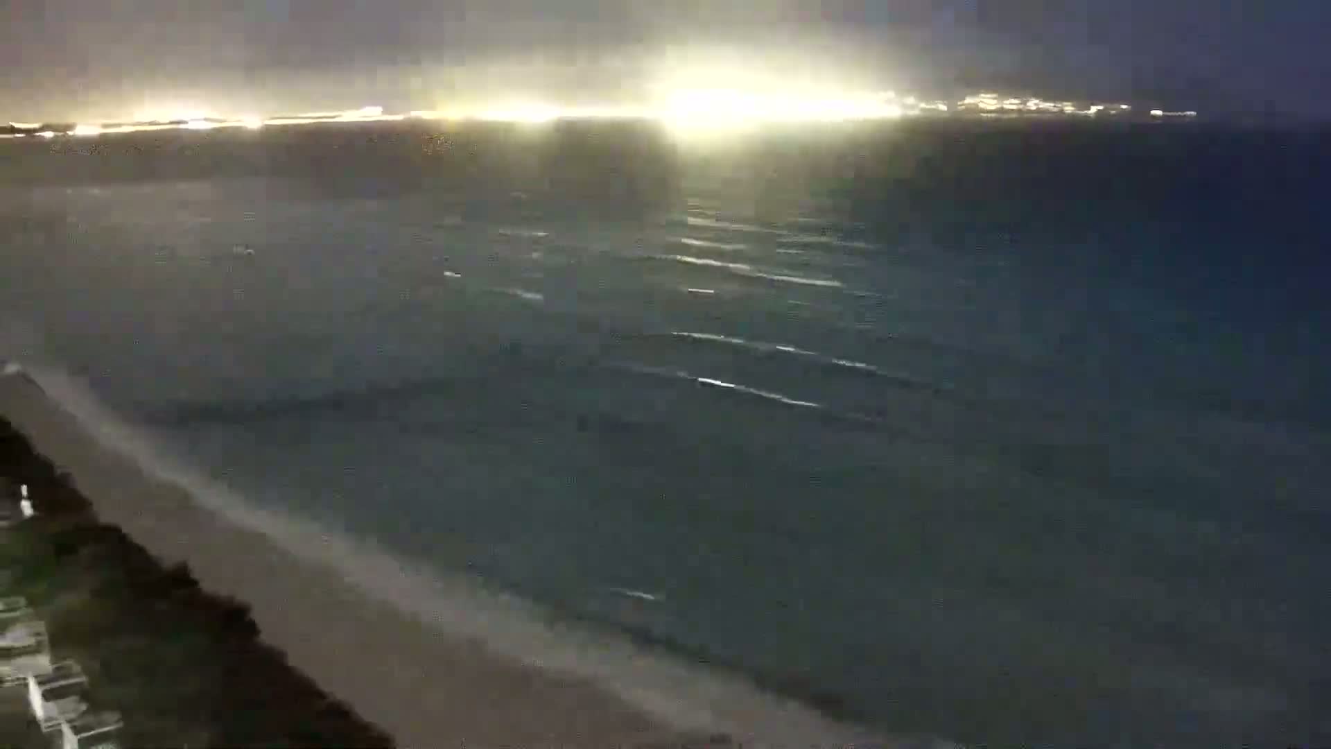 A nighttime view of a beach with gently lapping waves, illuminated by city lights in the distance.