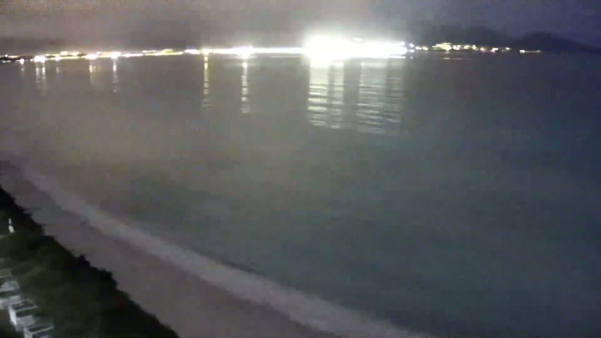 A nighttime view shows a calm, dark-colored body of water with a sandy beach and a distant shoreline illuminated by lights.