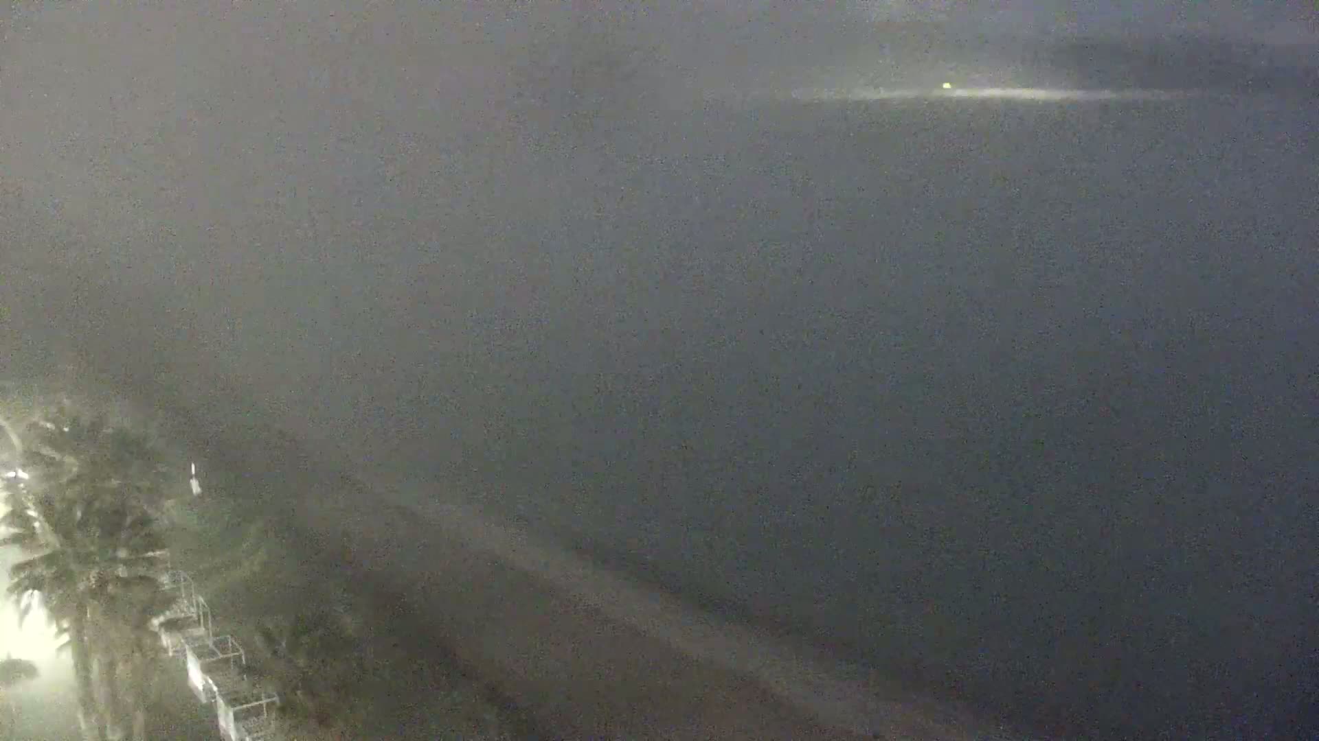 A nighttime view shows a hazy, dark ocean meeting a beach with palm trees and a structure, under a heavy fog or mist.