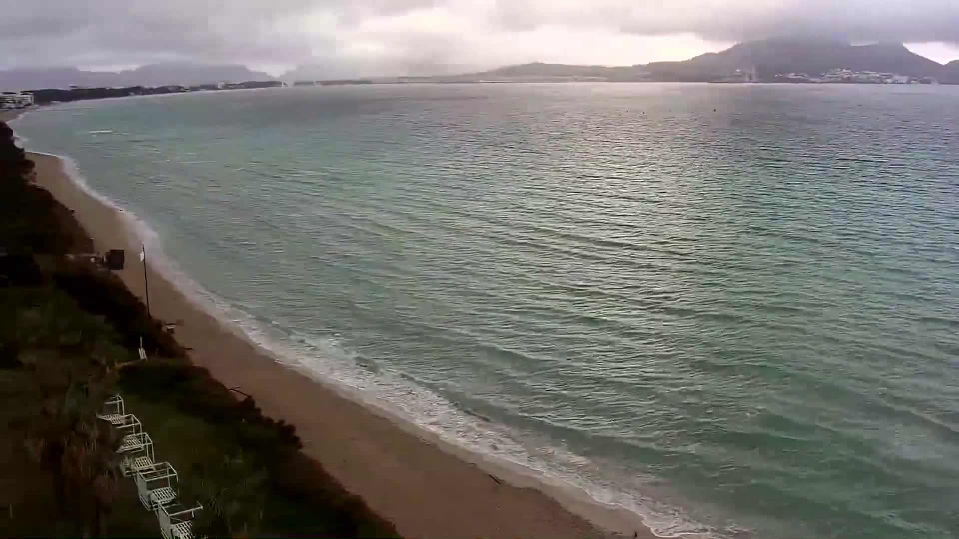 A sandy beach with gentle waves laps the shore under a cloudy sky, with calm water extending to distant land.