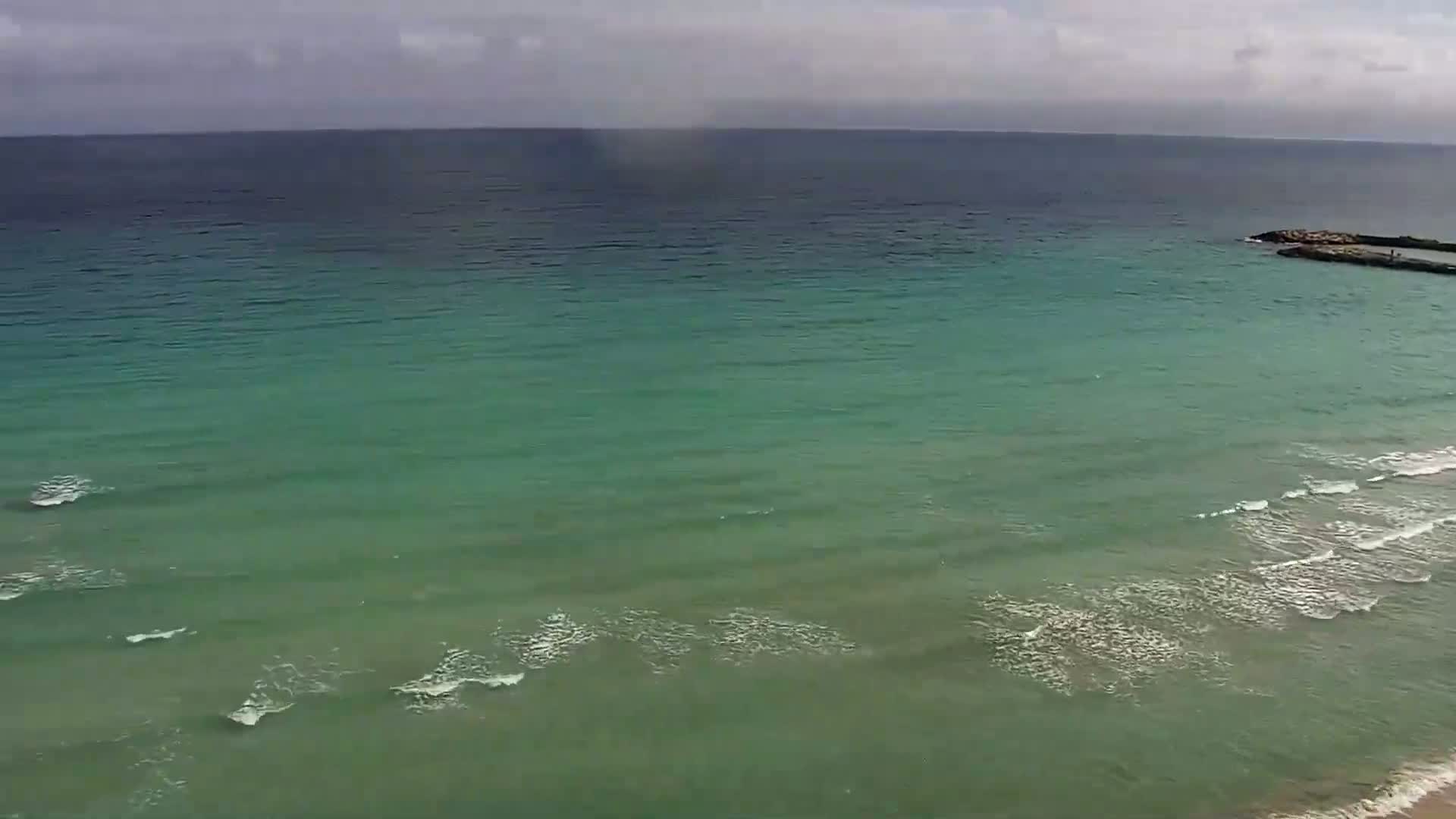 An aerial view shows a calm, teal-green ocean with small waves lapping the shore under a cloudy sky.