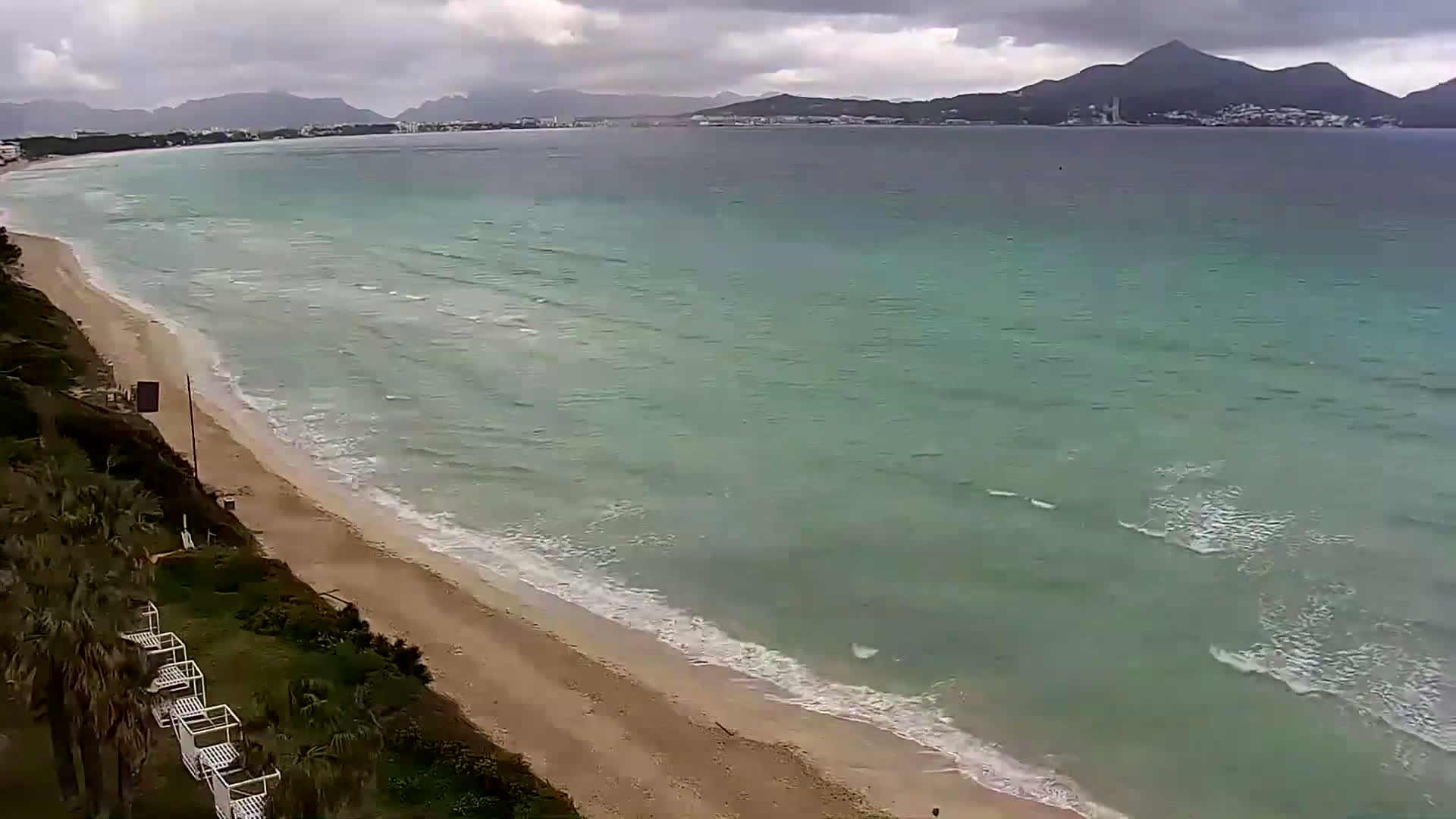 A sandy beach with gentle waves under a cloudy sky stretches towards a calm sea and distant mountains.