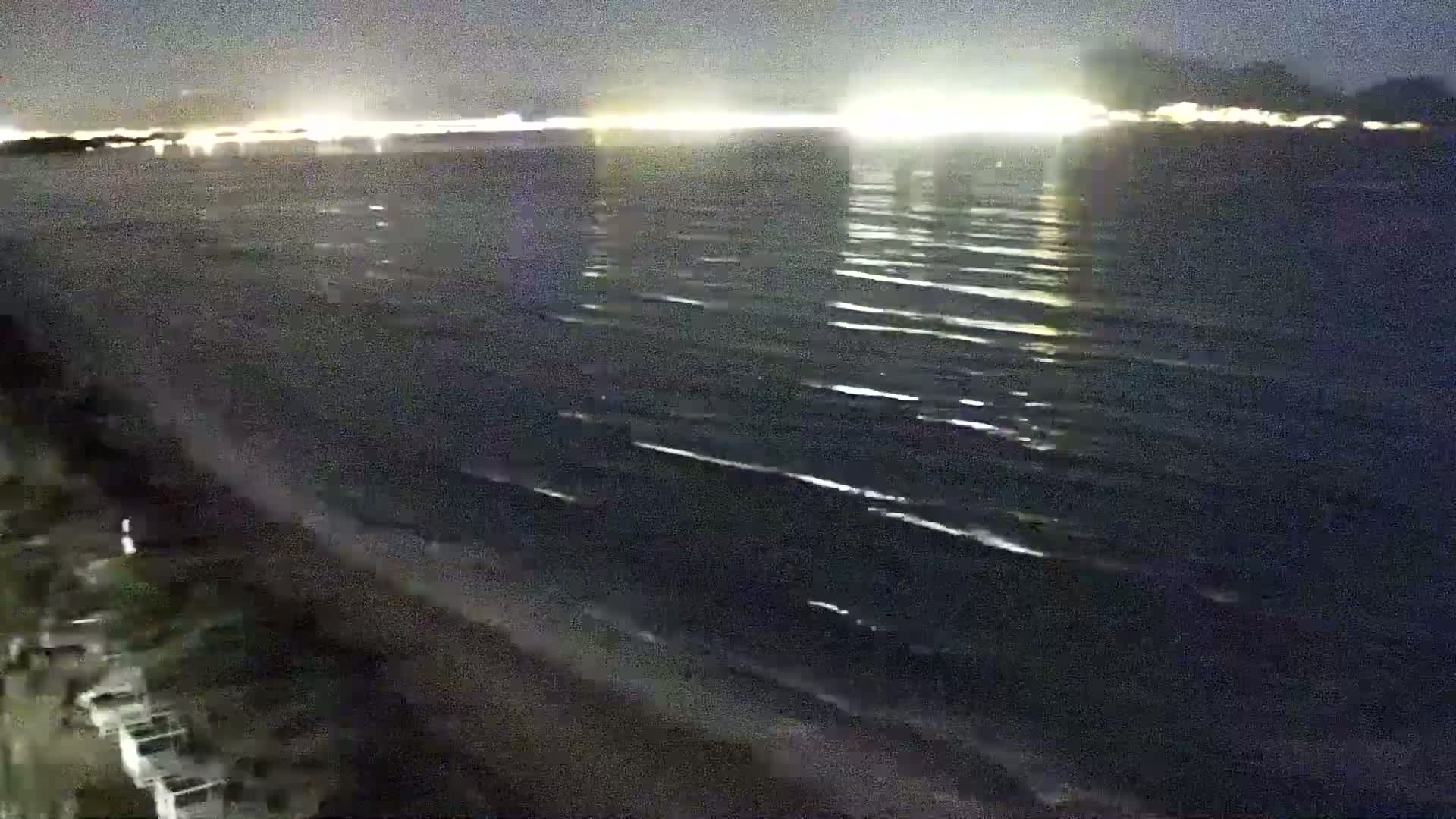 A dark, nighttime beach scene shows a gently waving sea under a mostly clear sky, with a distant shoreline illuminated by lights.