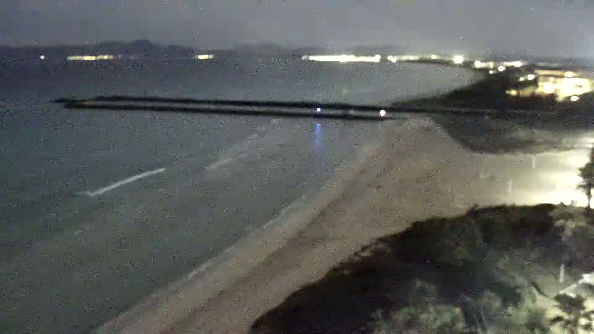 A nighttime, high-angle view shows a dark sandy beach with gentle waves lapping the shore, a breakwater in the water, and distant city lights across the bay.