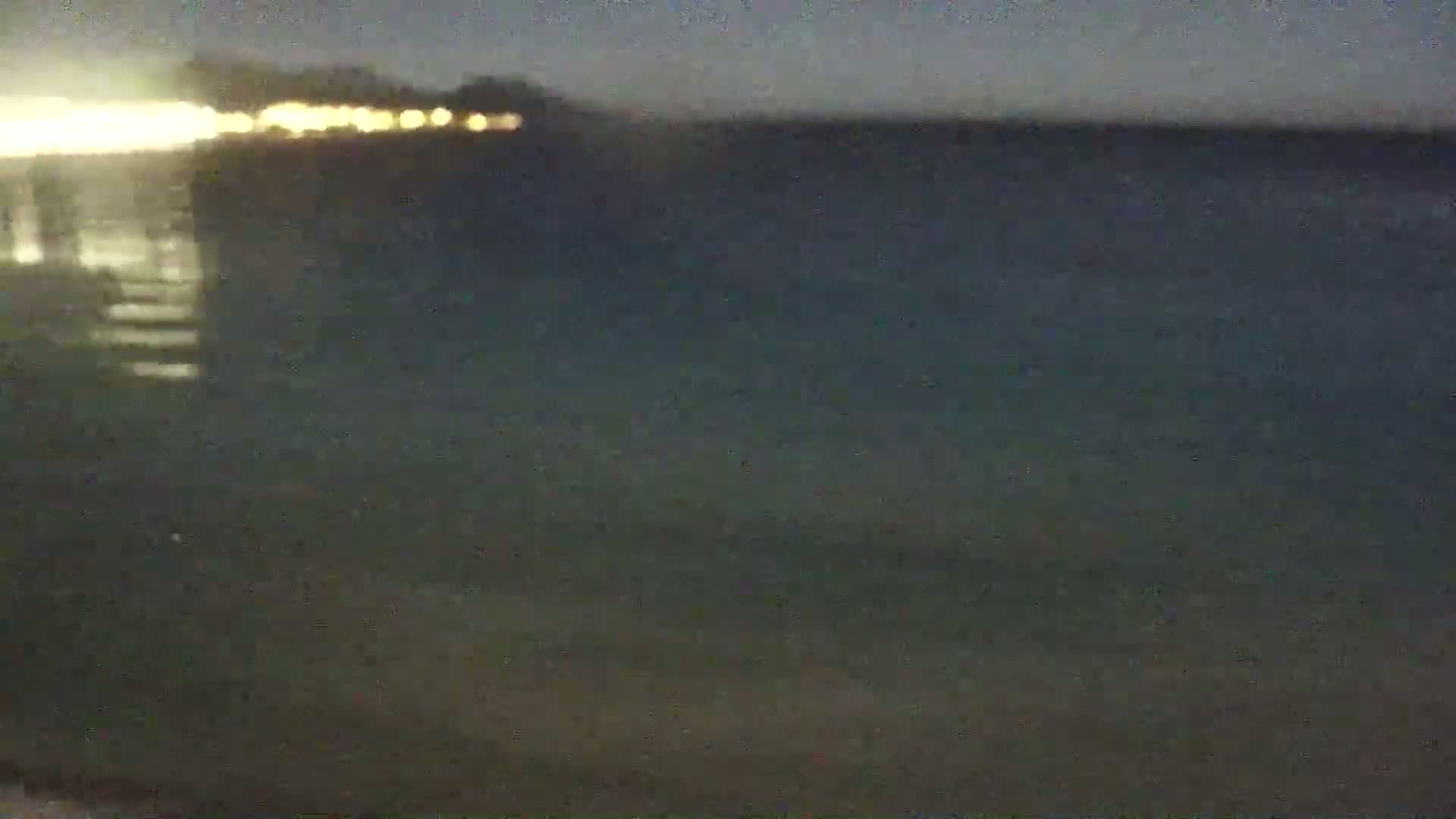 A blurry nighttime image shows a dark body of water with a faintly lit shoreline in the distance.