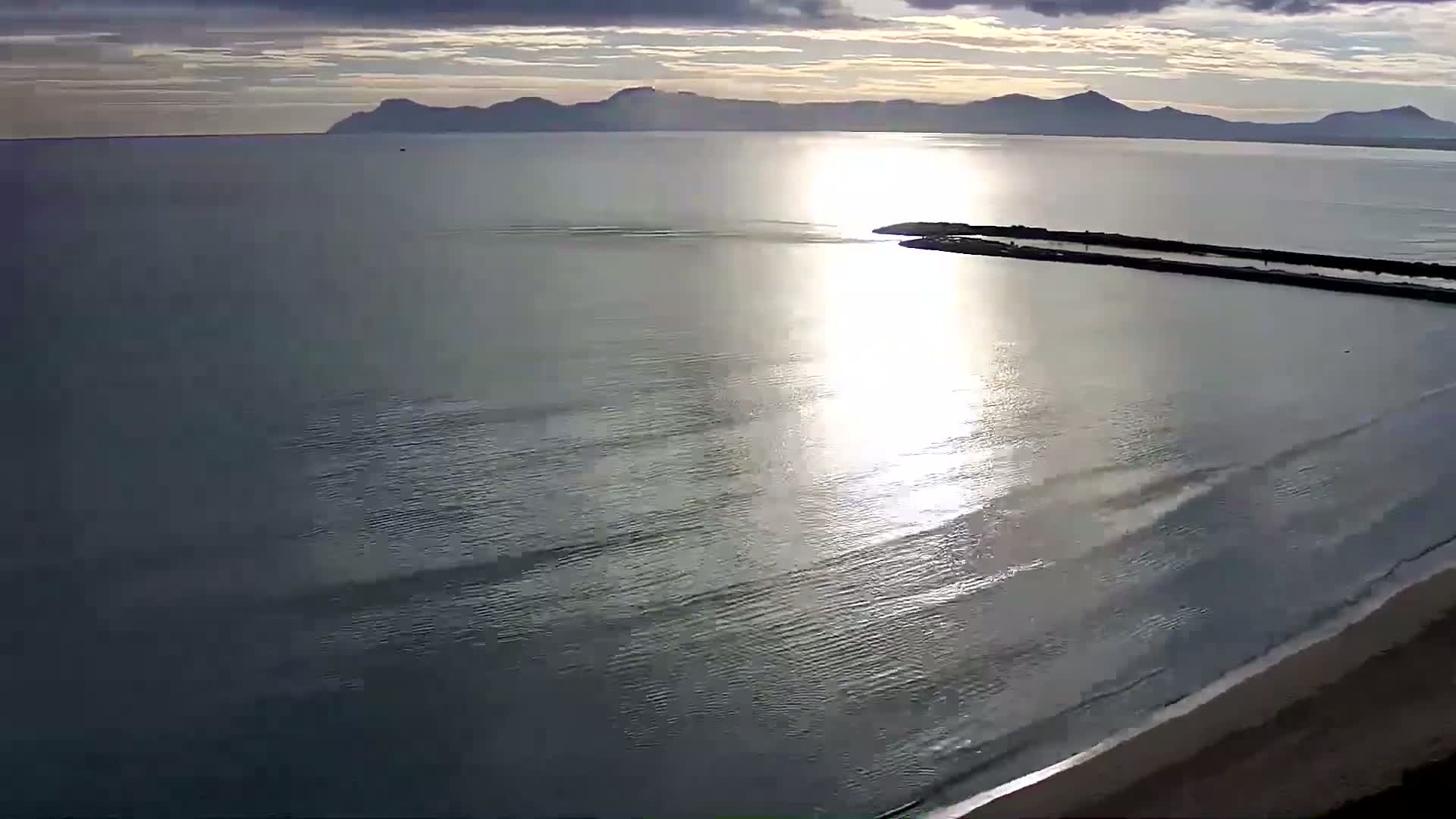 A calm, sunlit ocean meets a sandy beach, with a distant mountain range visible under a partly cloudy sky.