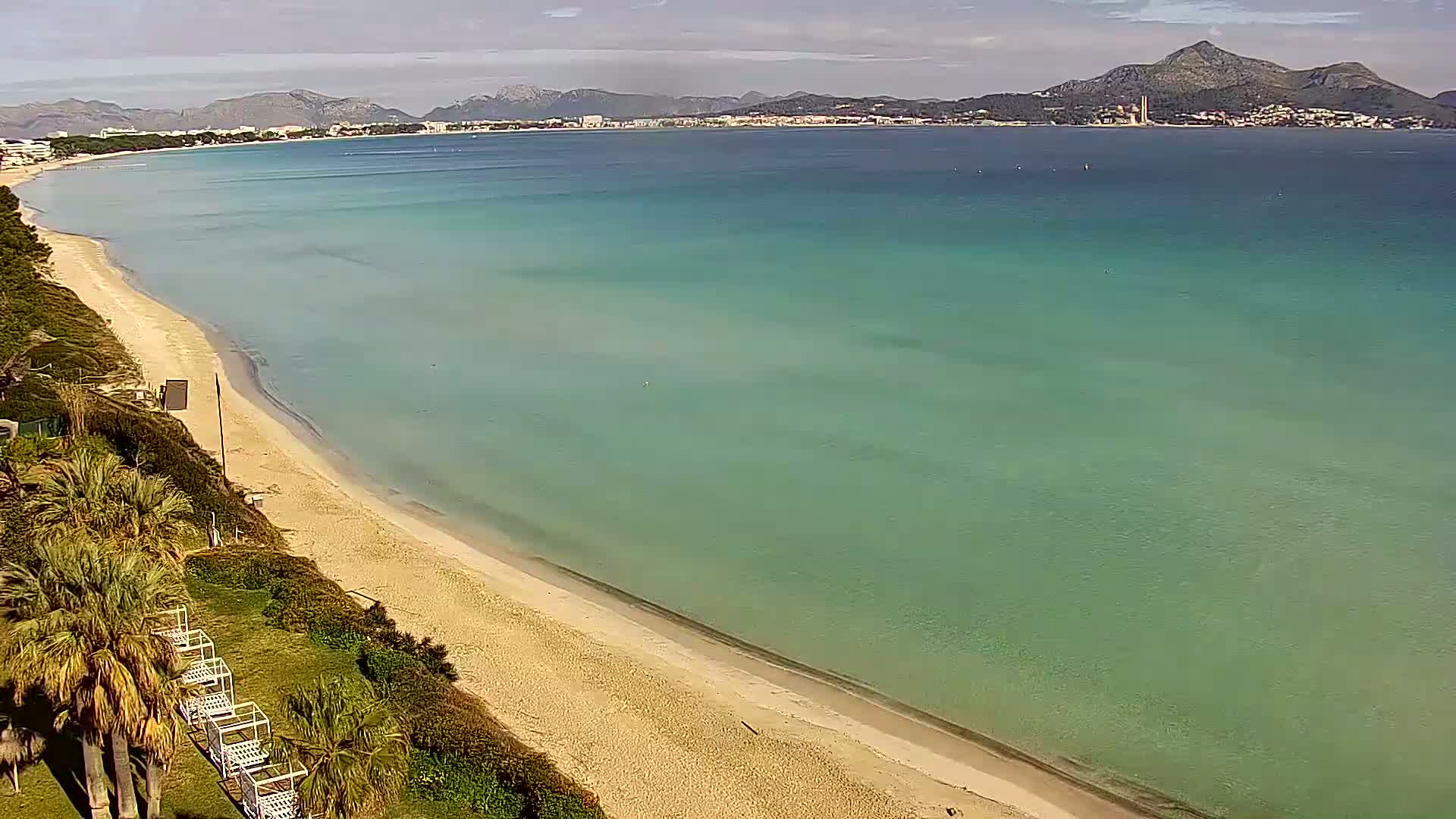 An aerial view shows a long sandy beach curving gently into a calm, clear turquoise sea under a mostly cloudy sky, with distant mountains visible in the background.